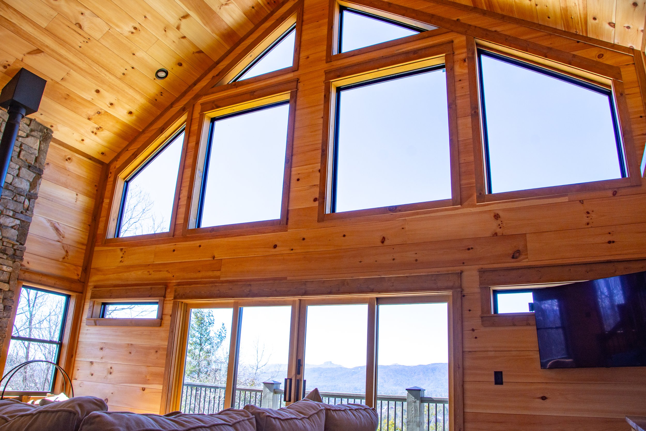 Interior of a wooden house with large, irregularly shaped windows showing a mountain landscape and blue sky outside.