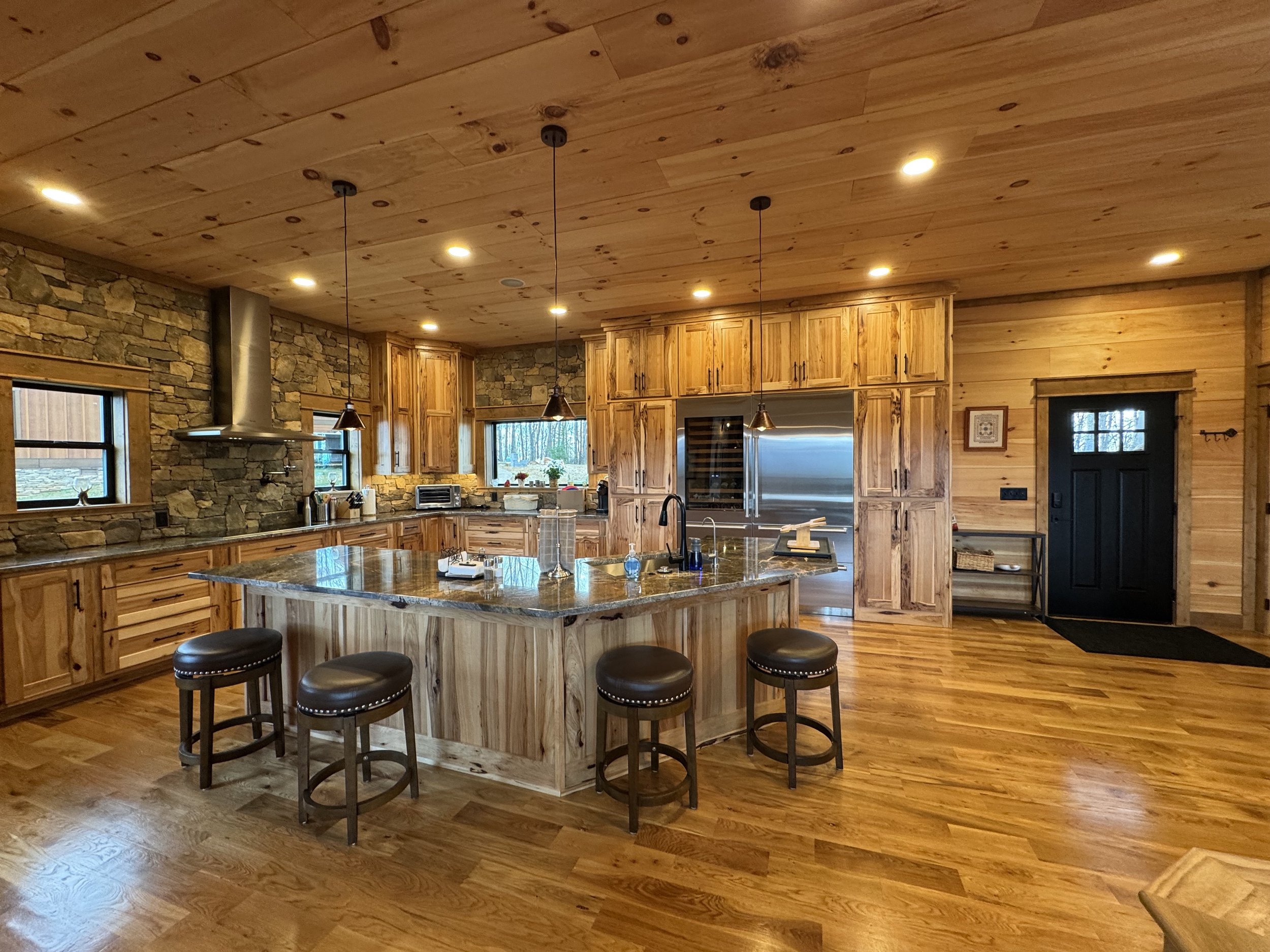 A spacious rustic kitchen with wooden cabinets, a large central island with a granite countertop, and a stainless steel refrigerator. The kitchen has a stone accent wall, large windows, and hardwood floors. There are three black barstools at the isla