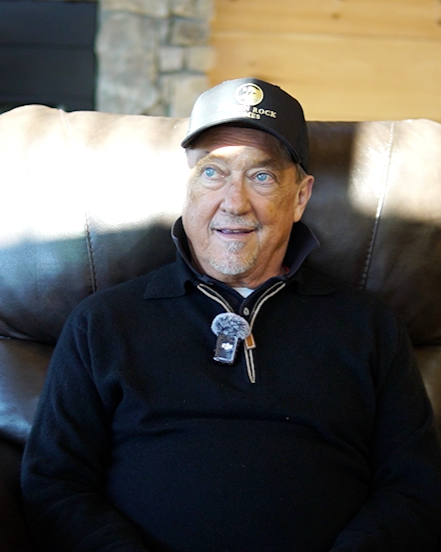 Man sitting on a leather couch wearing a black jacket, black cap, and a headset microphone, with a stone wall and wooden panel background.