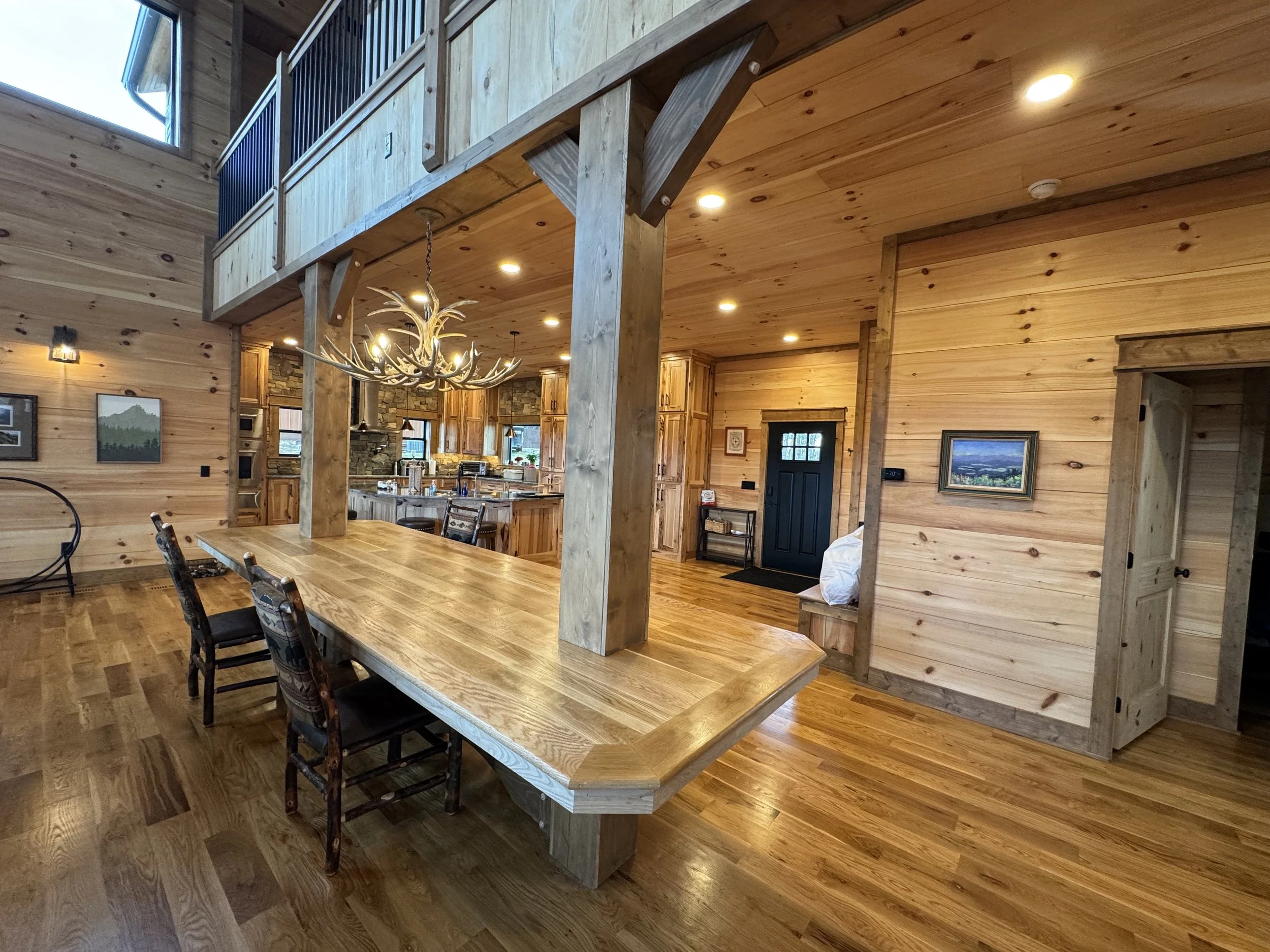 Interior of a wooden cabin-style home with an open kitchen and dining area. The room features wooden walls, floors, and furniture, including a large dining table with chairs. A chandelier resembling antlers hangs above the table, and a staircase lead