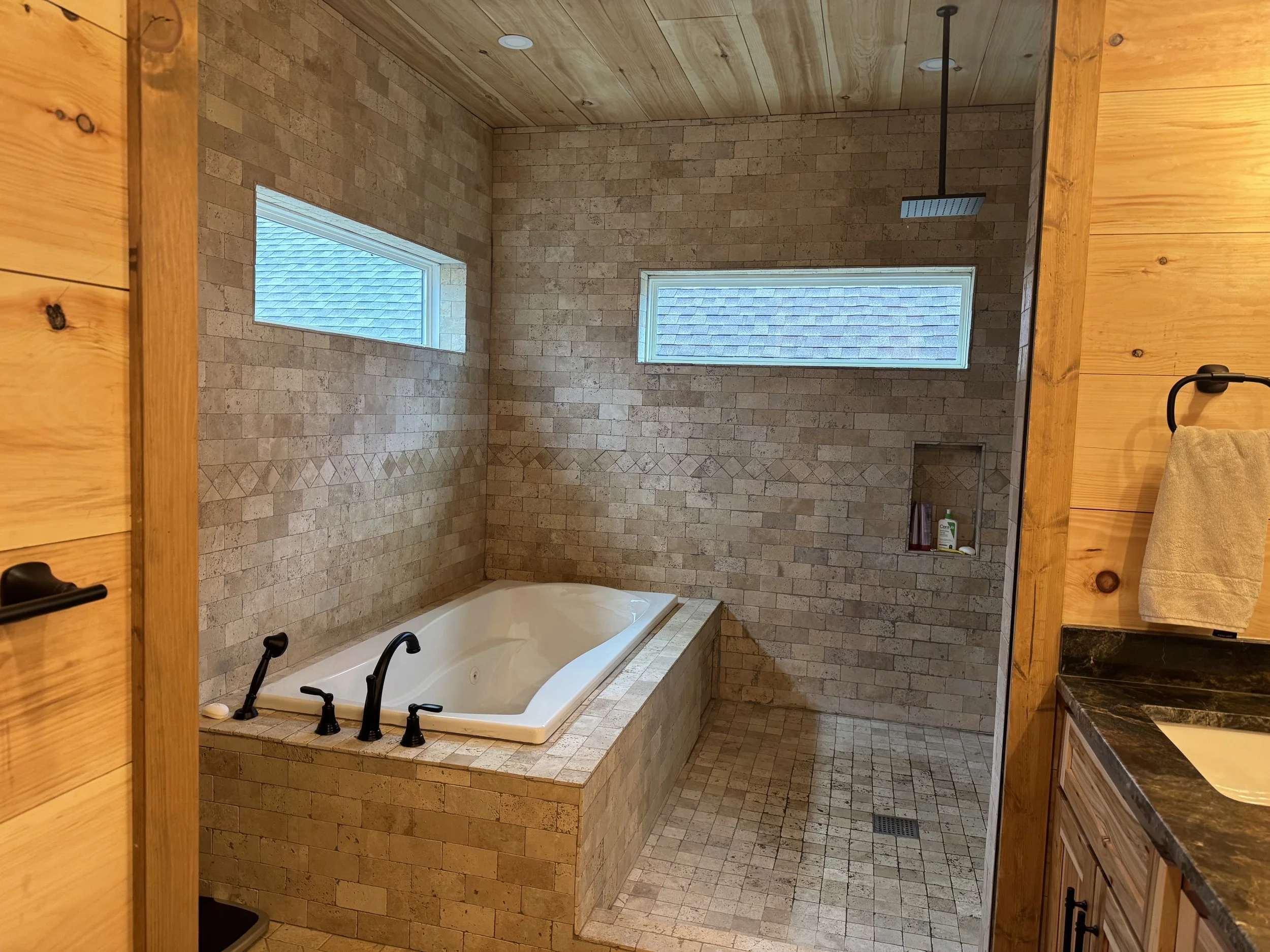 A bathroom with a large bathtub, beige brick tile walls, two horizontal windows, and a walk-in shower area with a wall niche and a rainfall showerhead.