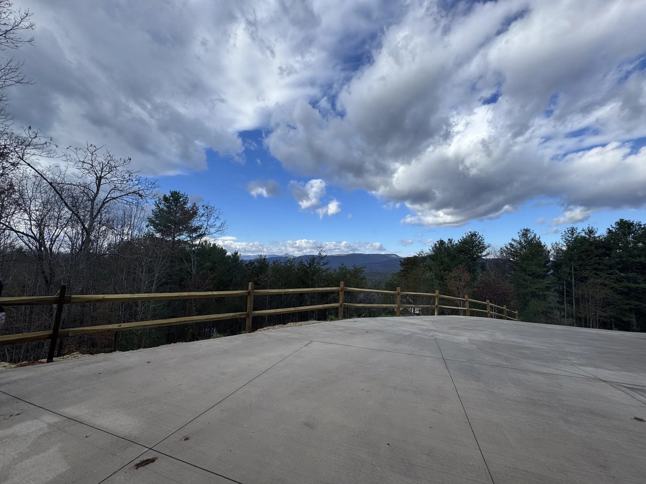 A wide view of a cloudy sky over a mountain range, with a concrete platform and a wooden fence in the foreground, surrounded by trees.