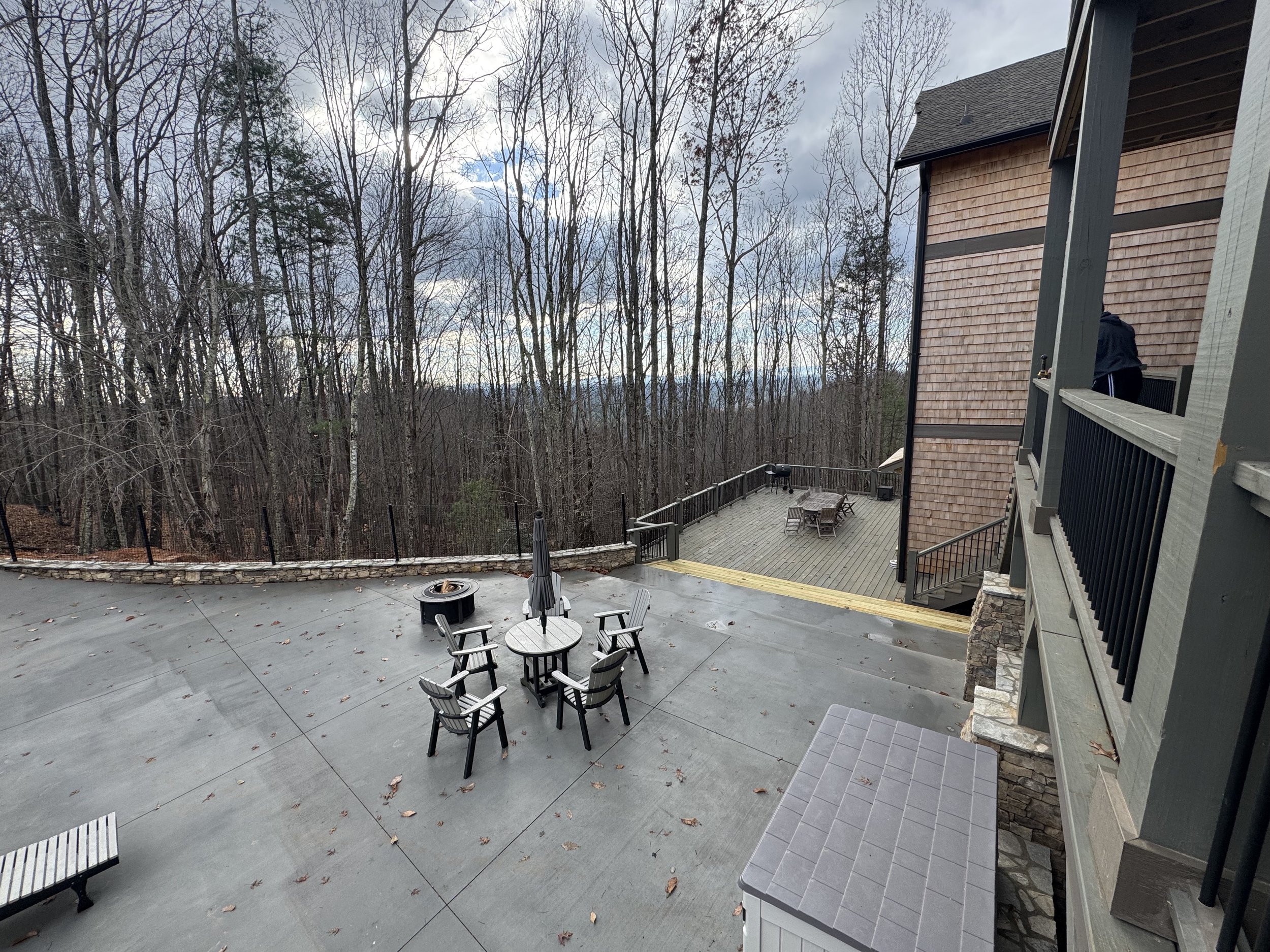 Outdoor patio area with a round table, six chairs, and a closed umbrella, surrounded by trees in a wooded landscape under an overcast sky.