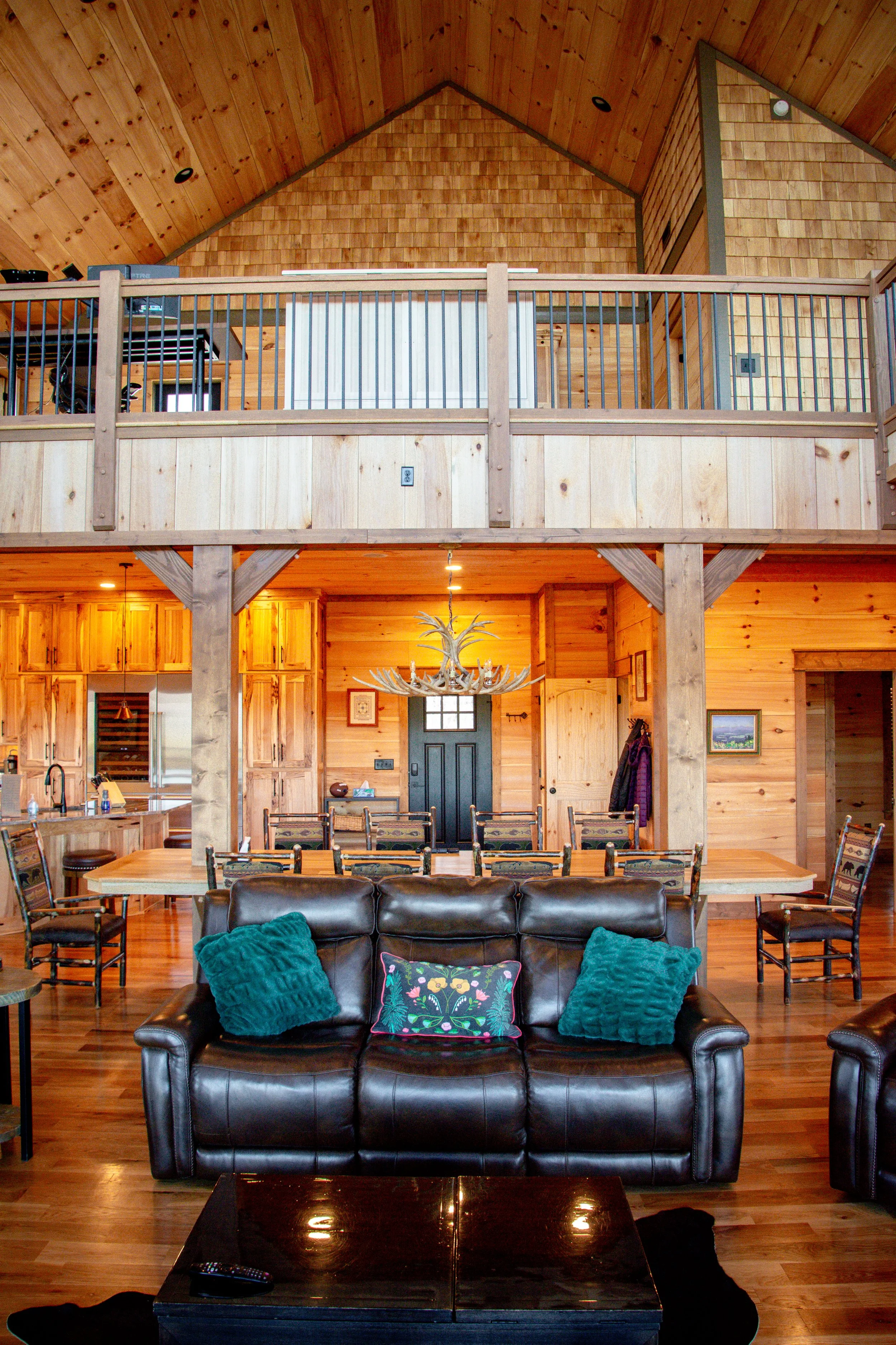 Interior of a wooden cabin with high ceiling, showcasing a living space with a leather sofa, colorful pillows, a large dining table, and a kitchen with wooden cabinets. There is an upper loft with a railing, and the room is decorated with a chandelie