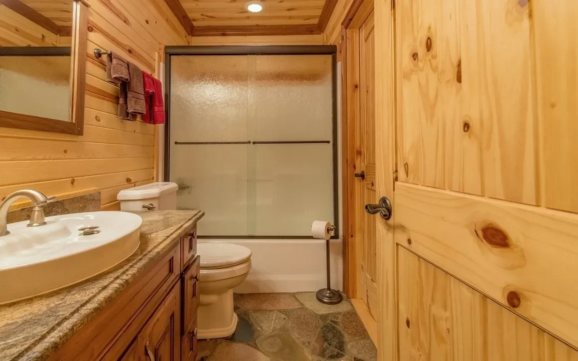 Small bathroom with wood-paneled walls, a sink with a granite countertop, a toilet, a shower with frosted sliding doors, a wall-mounted mirror, and a window seat with towels.