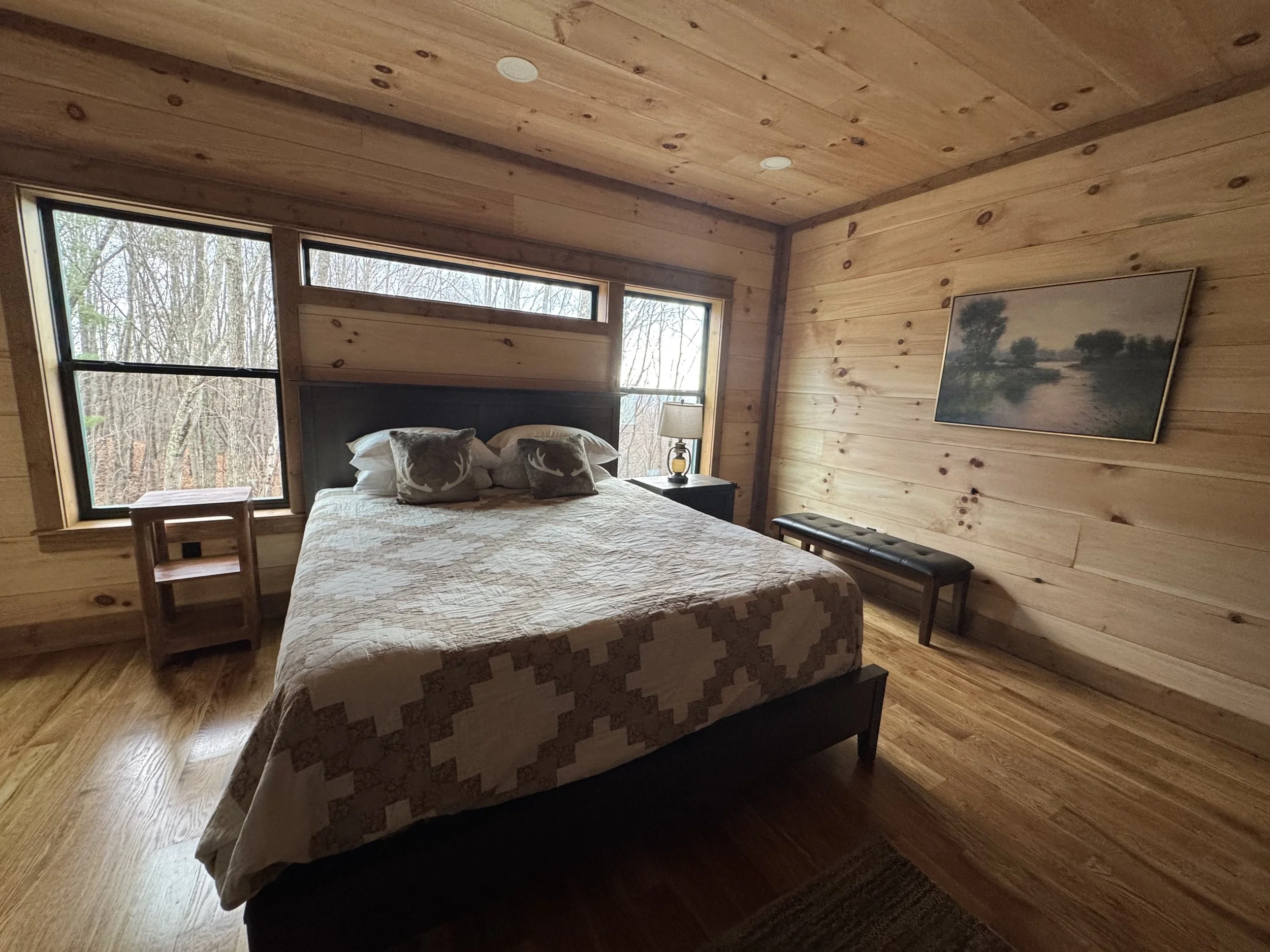 A cozy bedroom with wooden walls, a large bed with deer-themed pillows, a bedside table with a lamp, a window showing trees outside, a framed landscape painting on the wall, and a bench at the foot of the bed.