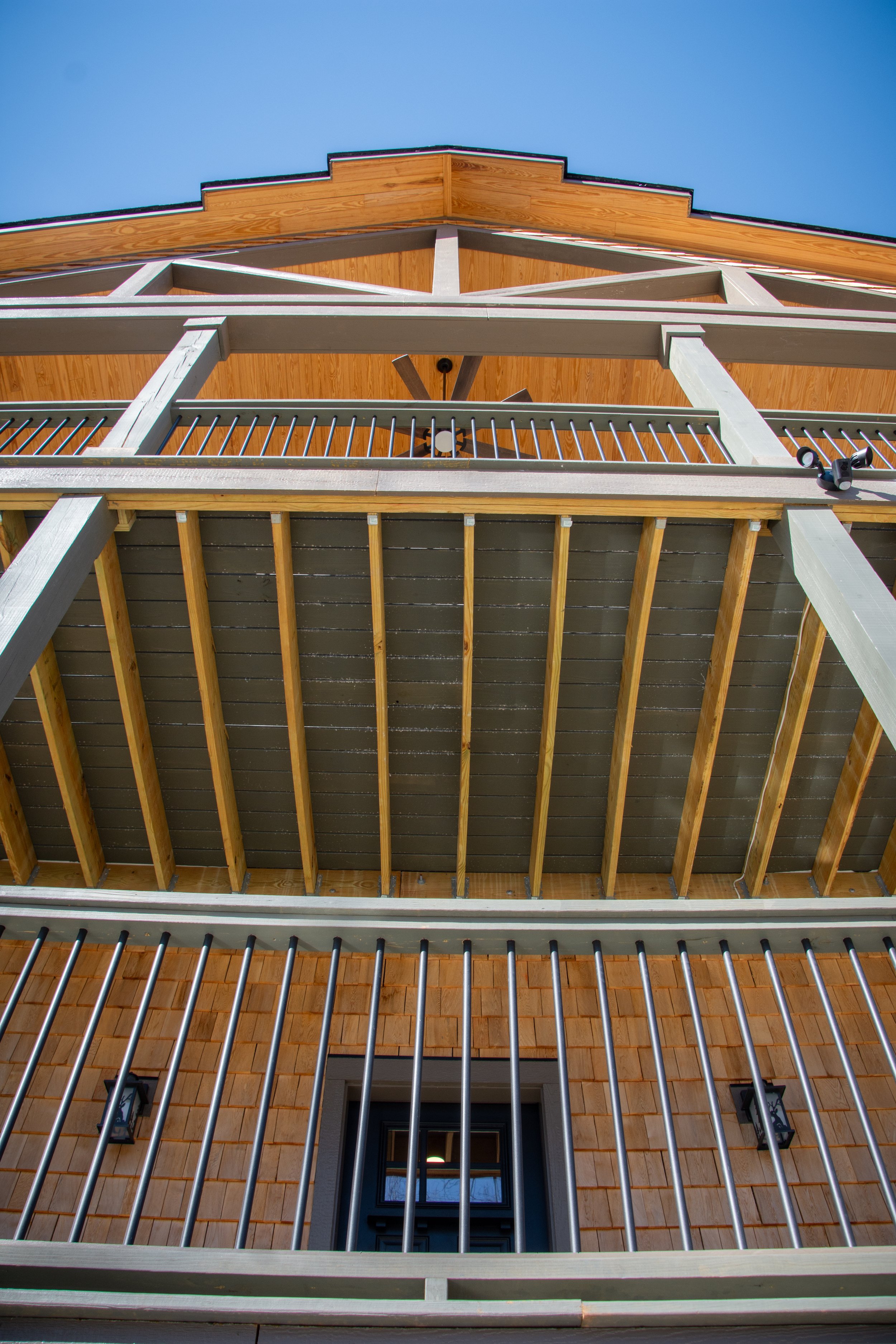 Looking up at a house's wooden balcony and roof from below, revealing wooden beams and metal railing, with a clear blue sky in the background.