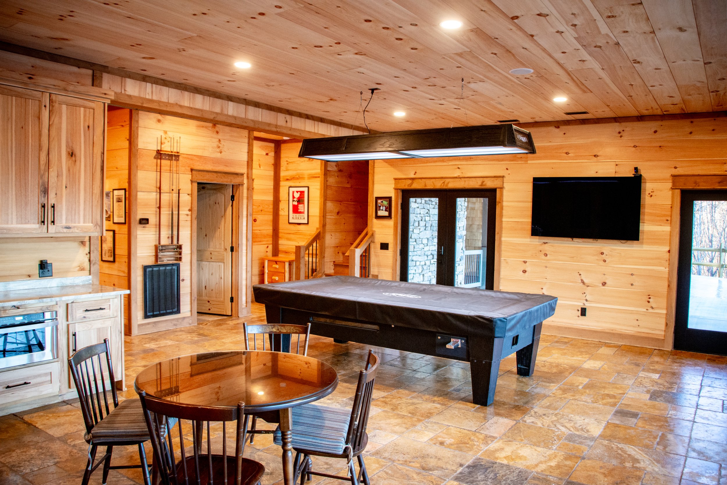Interior of a wood-paneled recreational room with a pool table, a flat-screen TV on the wall, a dining table with four chairs, and a sliding glass door leading outside.