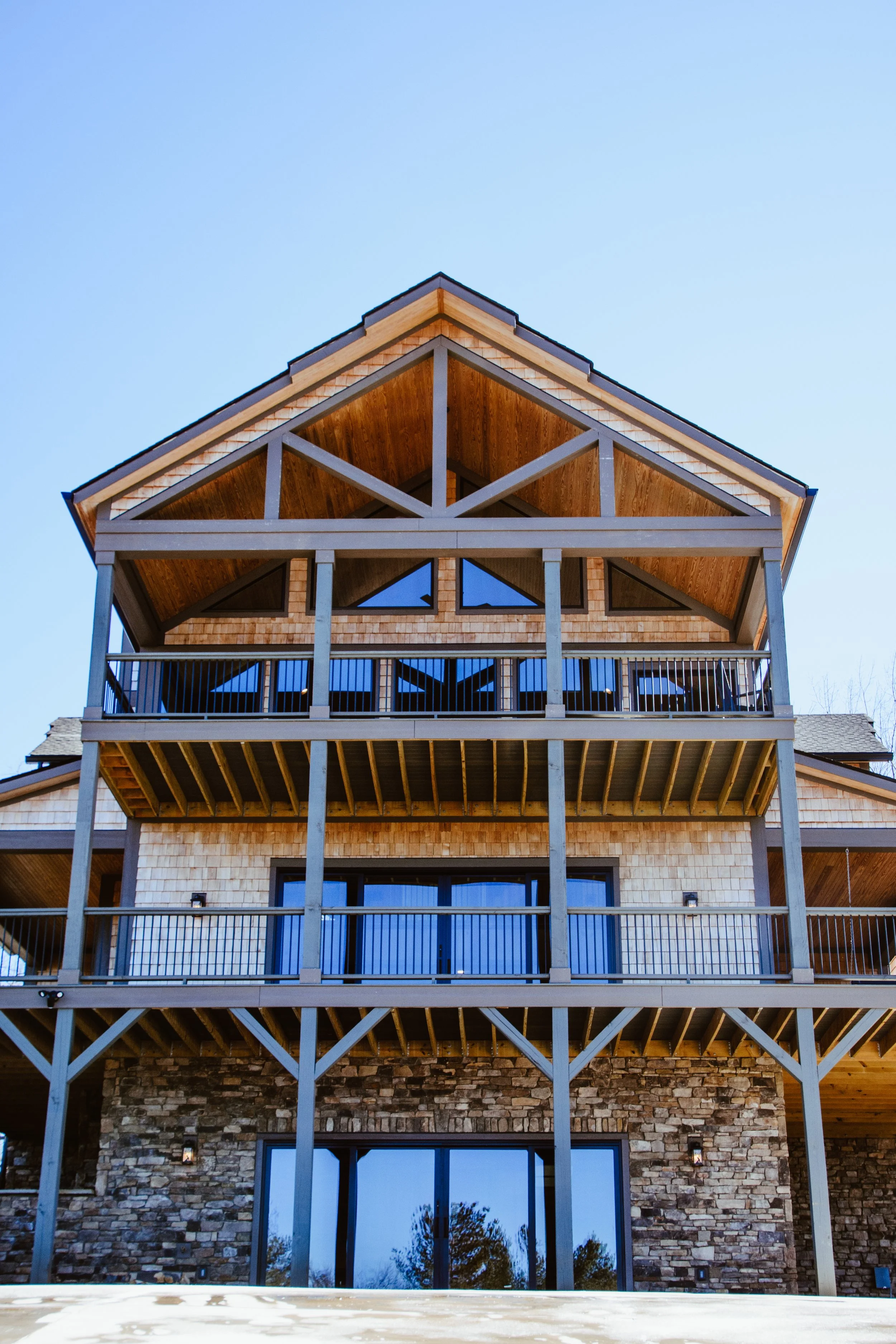 Front view of a multi-story house with a wooden and stone exterior, multiple balconies, and large glass doors, set against a clear blue sky.