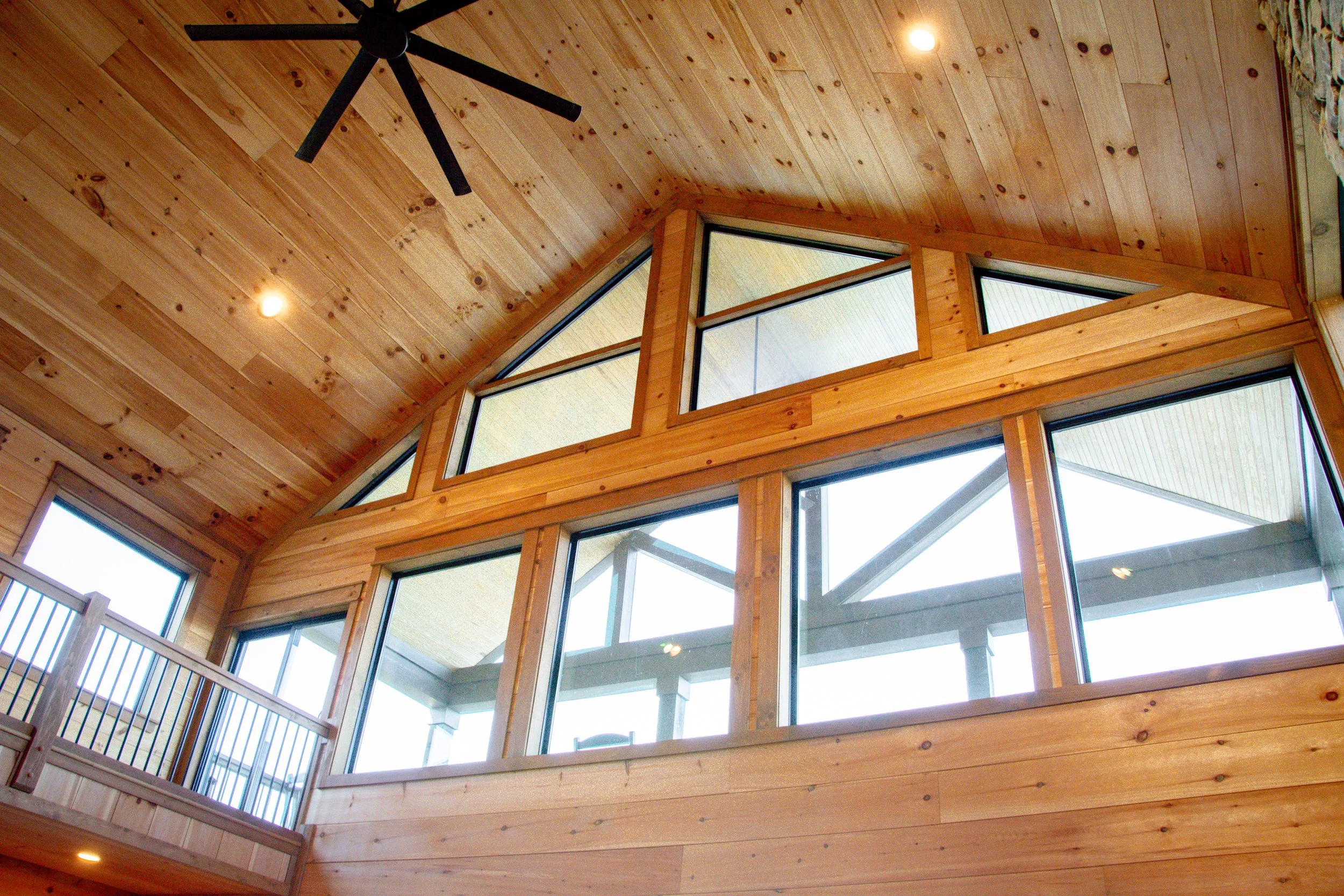 Interior view of a modern house with a tall wooden ceiling, large windows, and a ceiling fan.