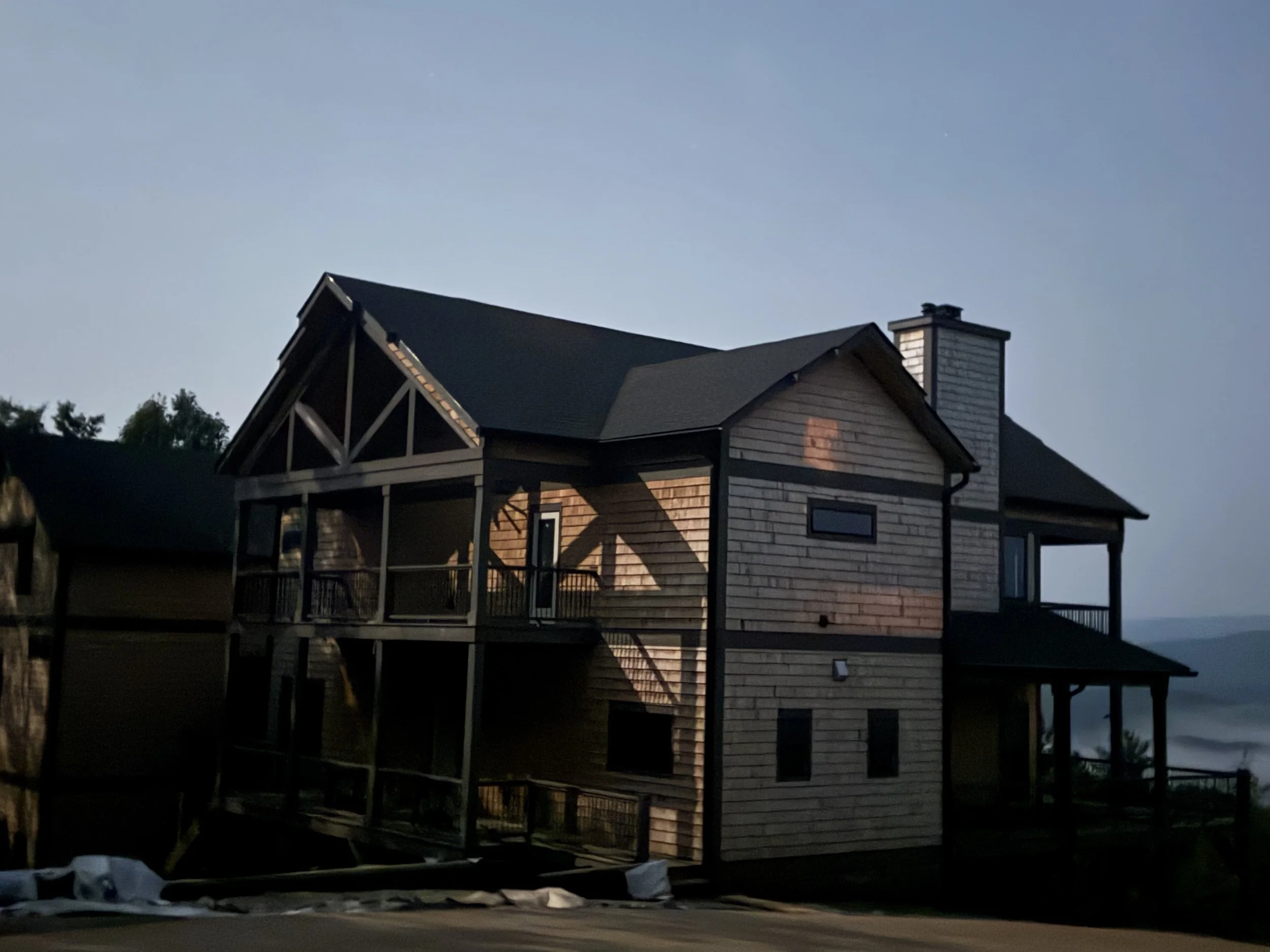 A large house with a sloped roof, vinyl siding, and a chimney, illuminated by natural sunlight, with a deck on the upper floor and a fenced yard.