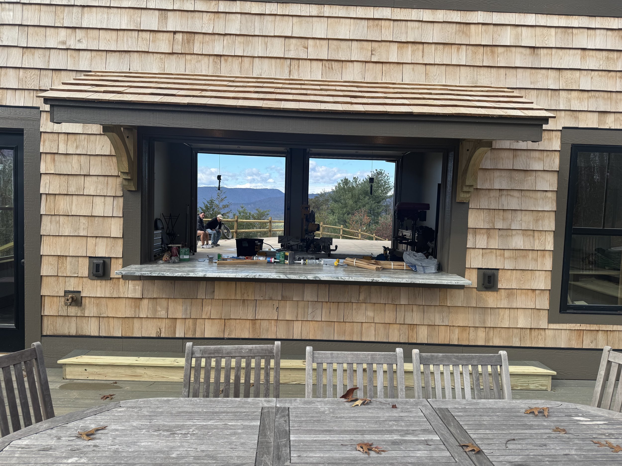 An outdoor wooden patio with a table and chairs in the foreground. The background features a house window with a view of mountains and trees. Inside the window, there are tools and objects, possibly indicating ongoing work or a renovation.