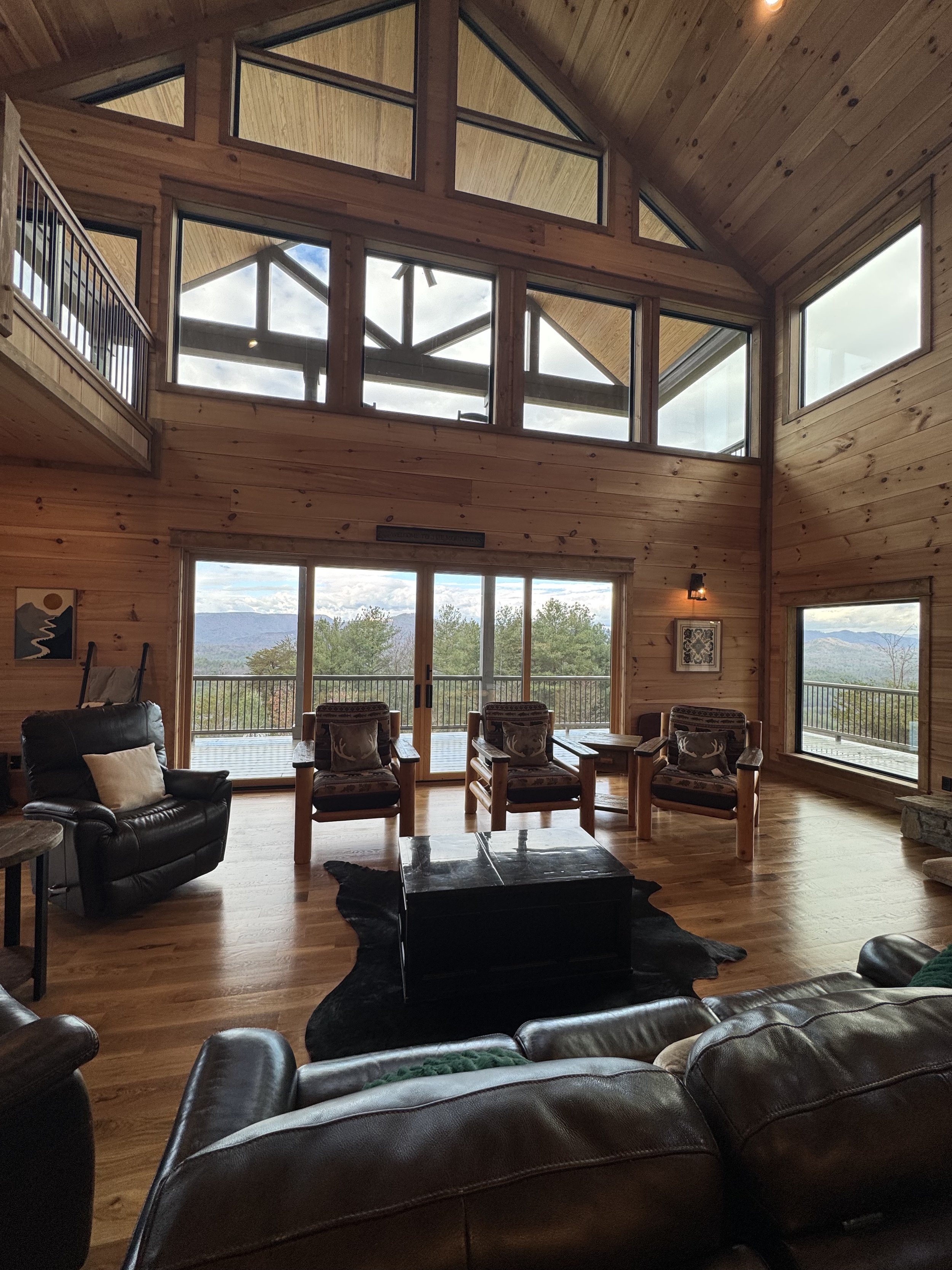 Interior view of a spacious living room with wooden walls and ceiling, featuring large windows and sliding glass doors opening to a balcony with a scenic mountain view. The room has leather and wooden chairs, a black coffee table on a black fur rug, 