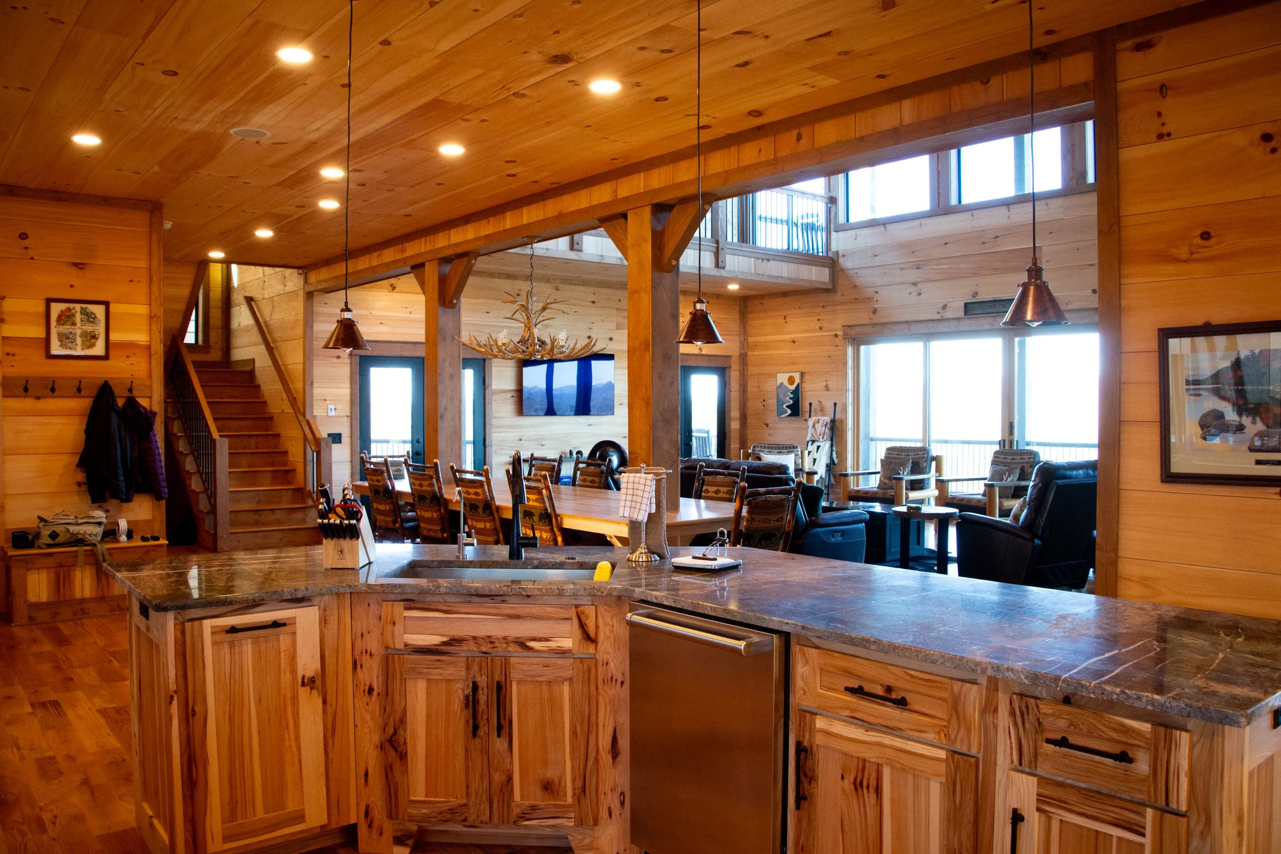 Interior of a rustic-style open-concept living space with wooden walls, ceiling, and furnishings, including a kitchen with a granite countertop, various chairs around a large dining table, seating area with armchairs and sofas, staircase, and large w