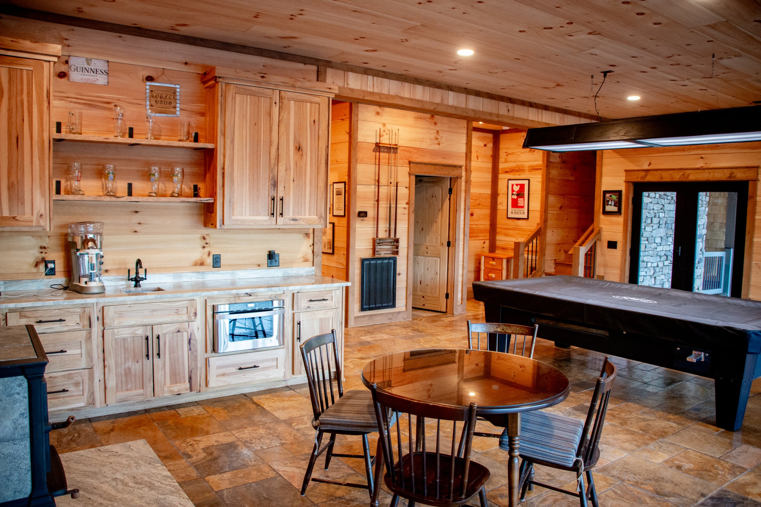 A rustic indoor room with wooden walls, ceiling, and cabinets, featuring a small kitchen area, a round dining table with four chairs, an electronic dartboard, and a black pool table covered with a protective cloth.