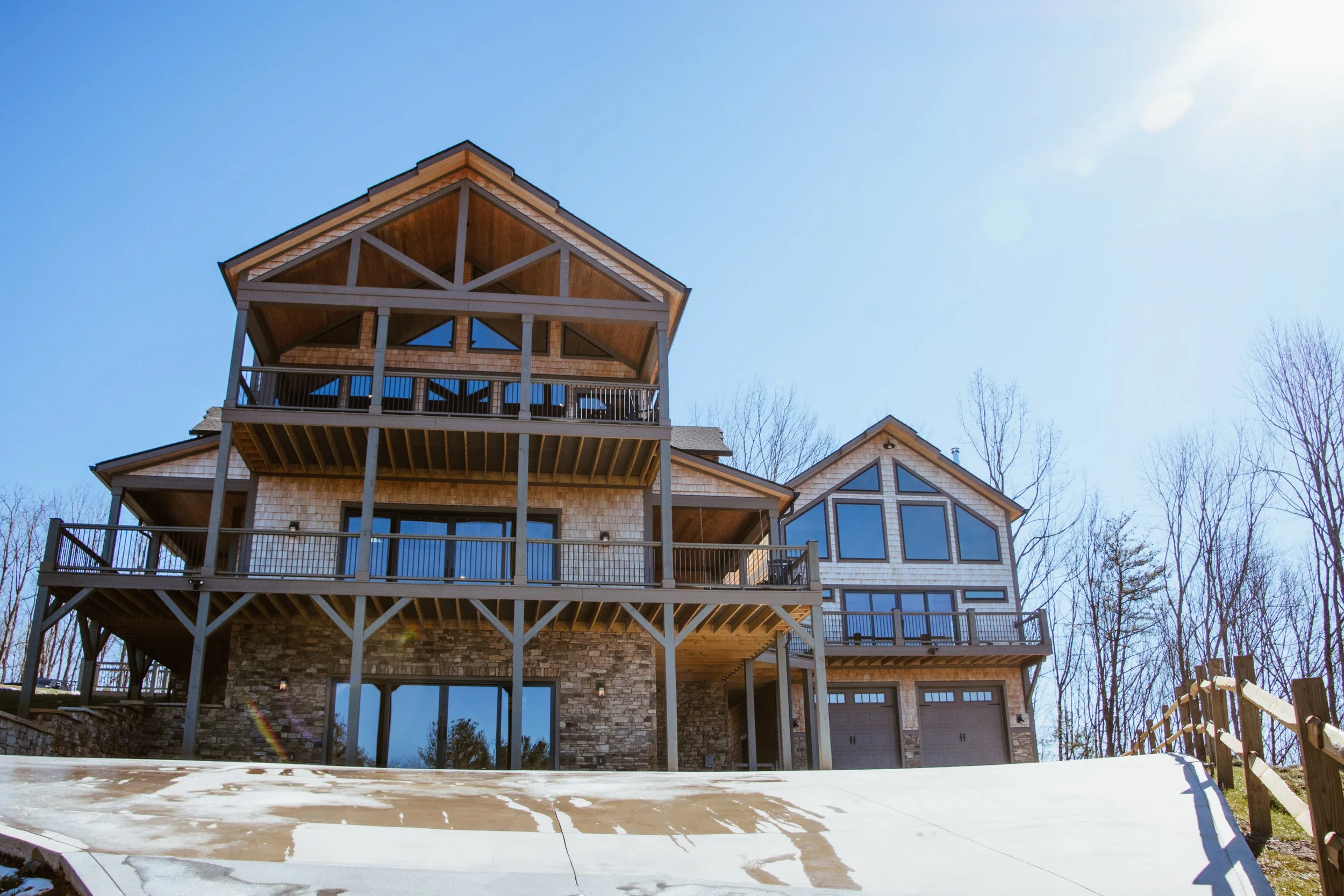 A large modern house with multiple levels, wooden balconies, large windows, stone accents, and a garage, set against a clear blue sky with bare trees in the background.