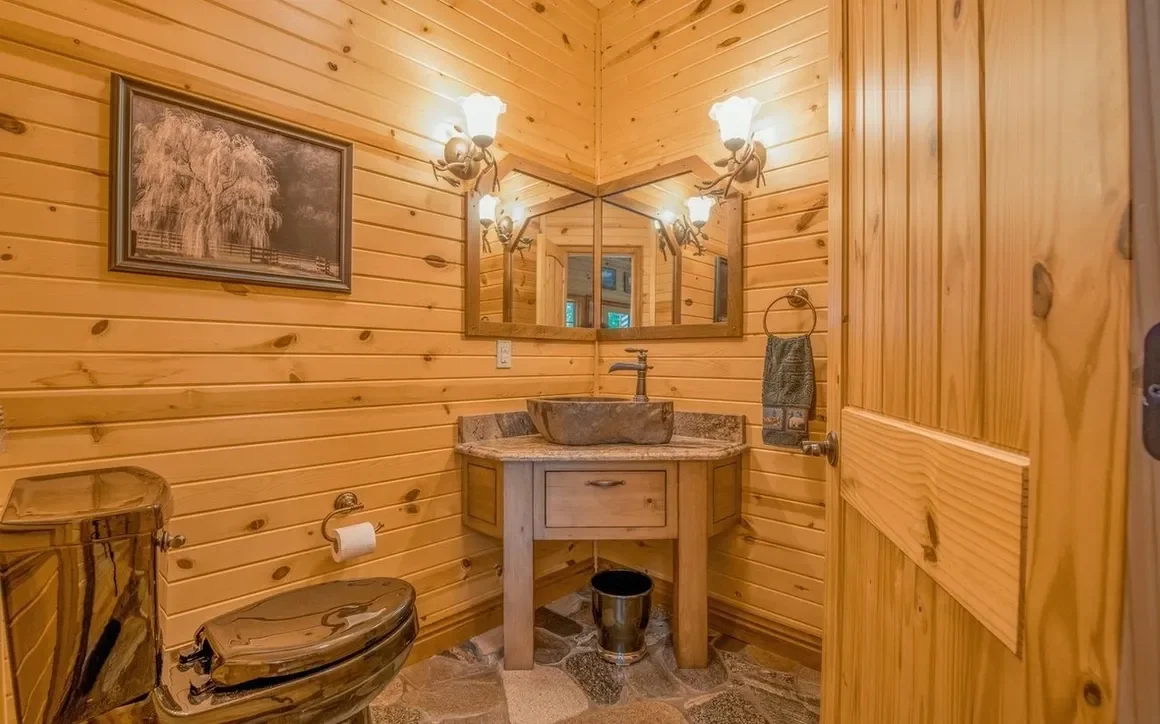 A rustic bathroom featuring wood-paneled walls, a stone vessel sink on a wooden vanity, a large mirror with a corner design, vintage wall sconces, a bronze toilet, and a framed black-and-white picture of a tree outside. The floor is made of stone til
