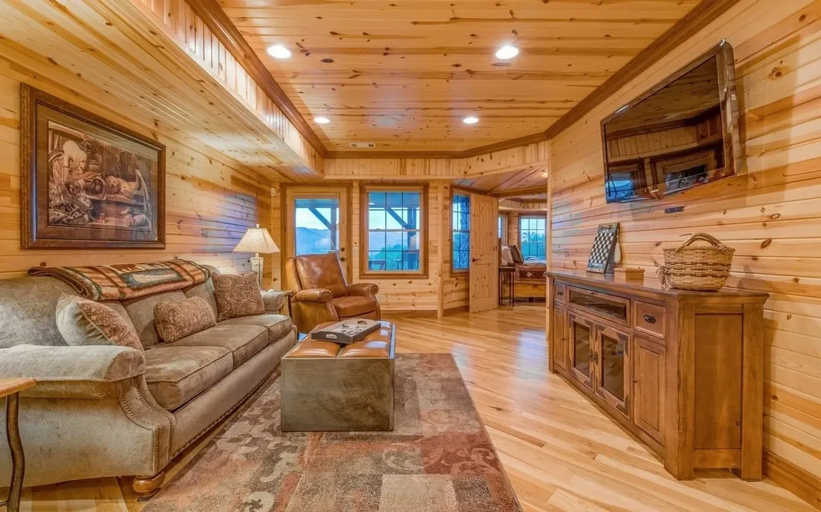 Cozy living room with wood-paneled walls and ceiling, featuring a beige sofa with pillows, a leather armchair, a wooden cabinet, a flat-screen TV, and windows showing an outdoor view.