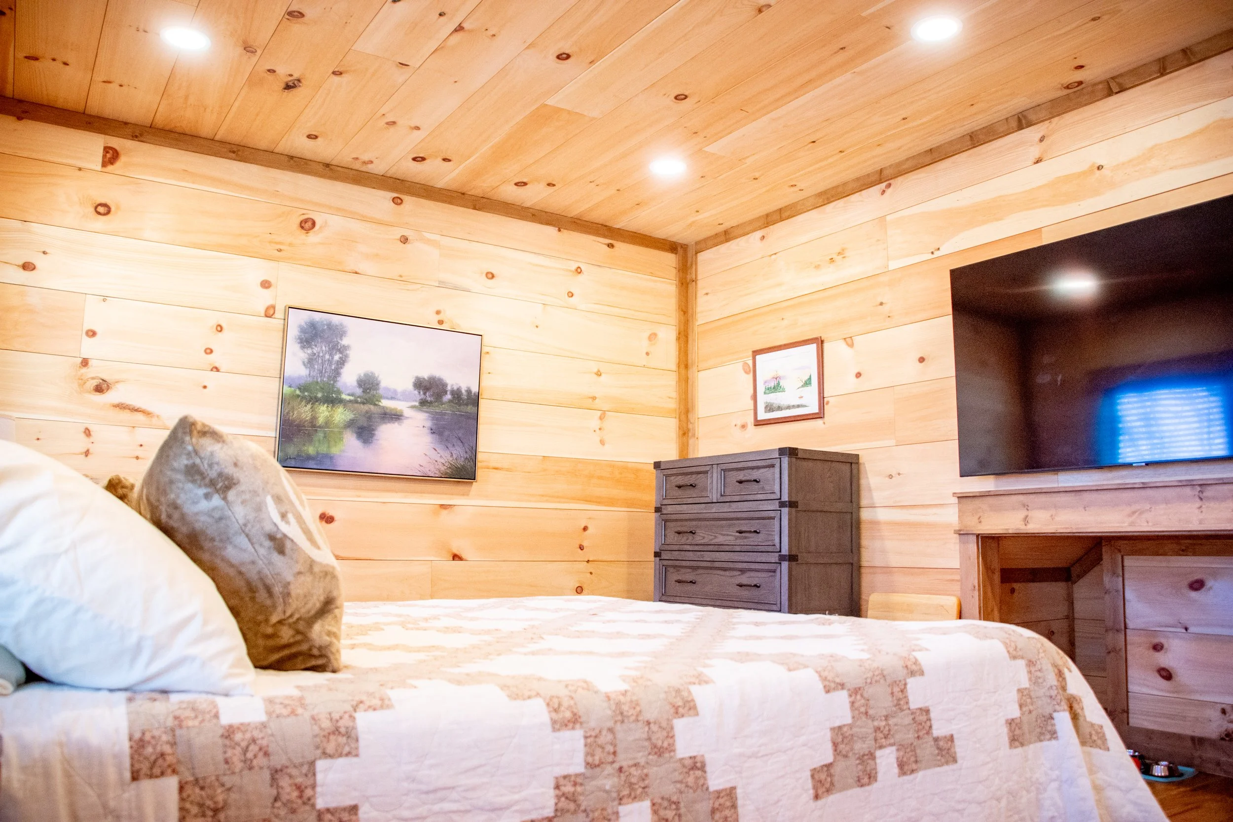 A bedroom with wooden paneled walls and ceiling, a bed with white and beige quilt, pillows, a gray chest of drawers, a framed painting of a river scene, and a large flat-screen TV mounted on the wall.