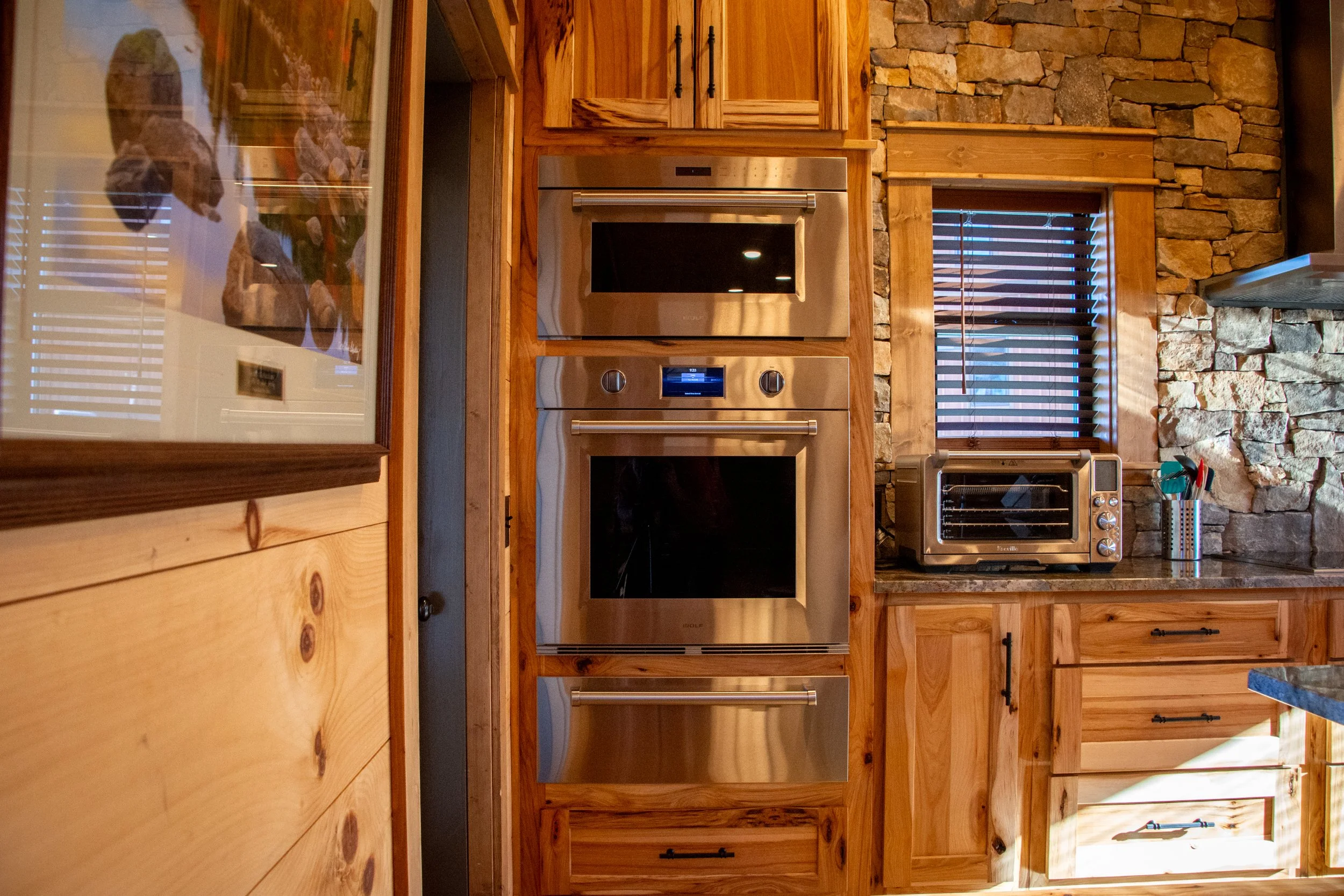 Kitchen with stainless steel oven and microwave, wooden cabinetry, stone wall, window with blinds, toaster oven, and countertop utensil holder.