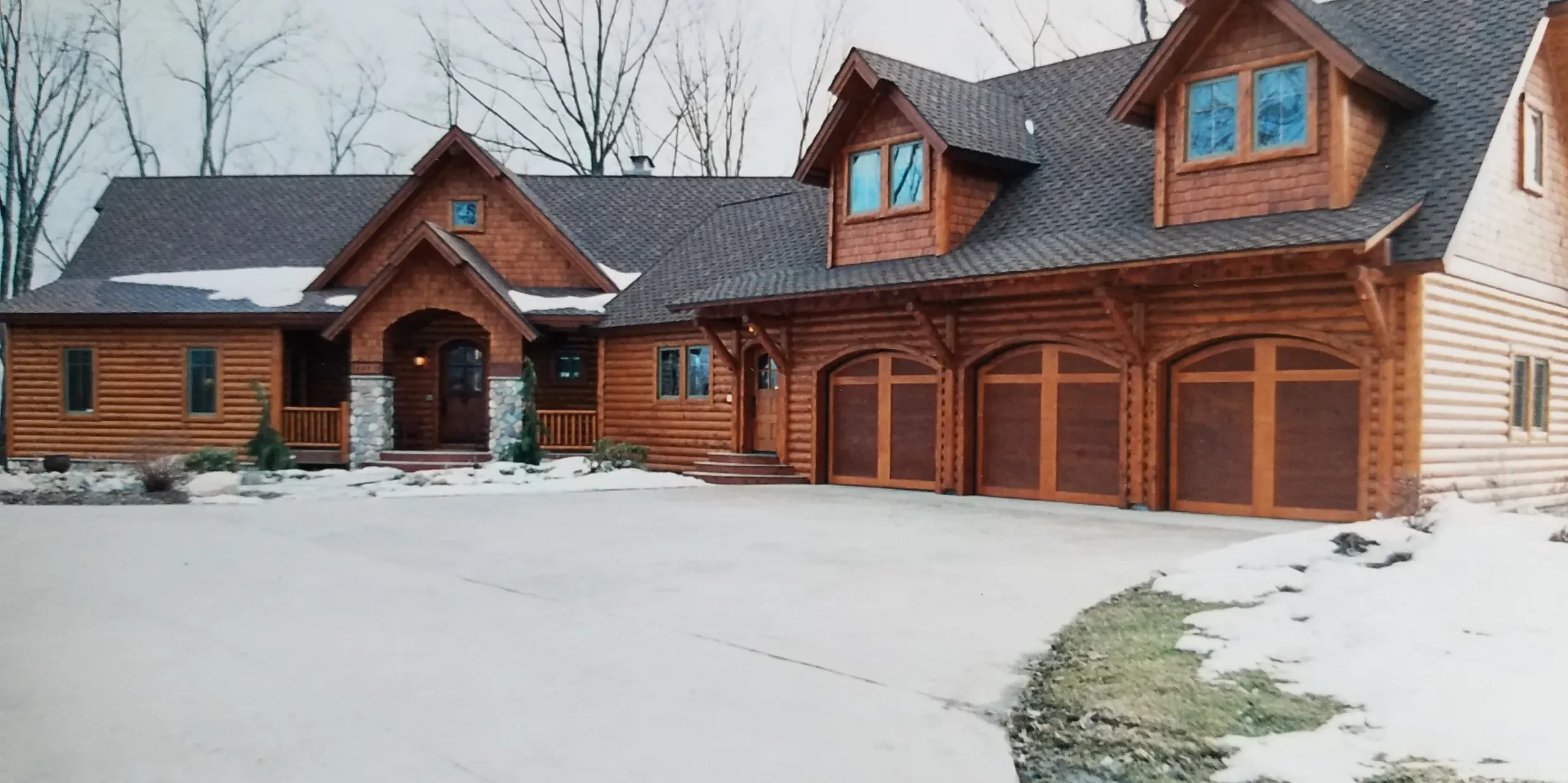 A large wooden lodge-style house with multiple gables, stone accents, and three garage doors, surrounded by snow and leafless trees.