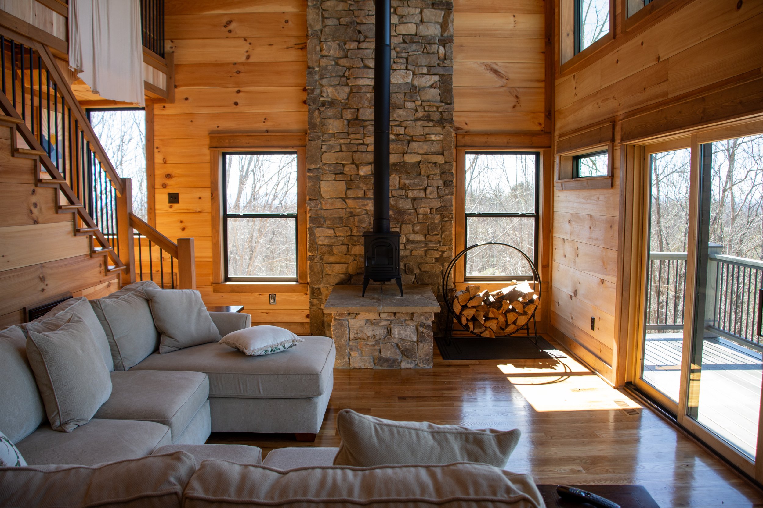 Living room with wooden walls, a stone fireplace with a stove pipe, a beige sectional sofa, windows showing a wooded outdoor view, a woodpile in a metal holder, and a sliding glass door to a balcony.