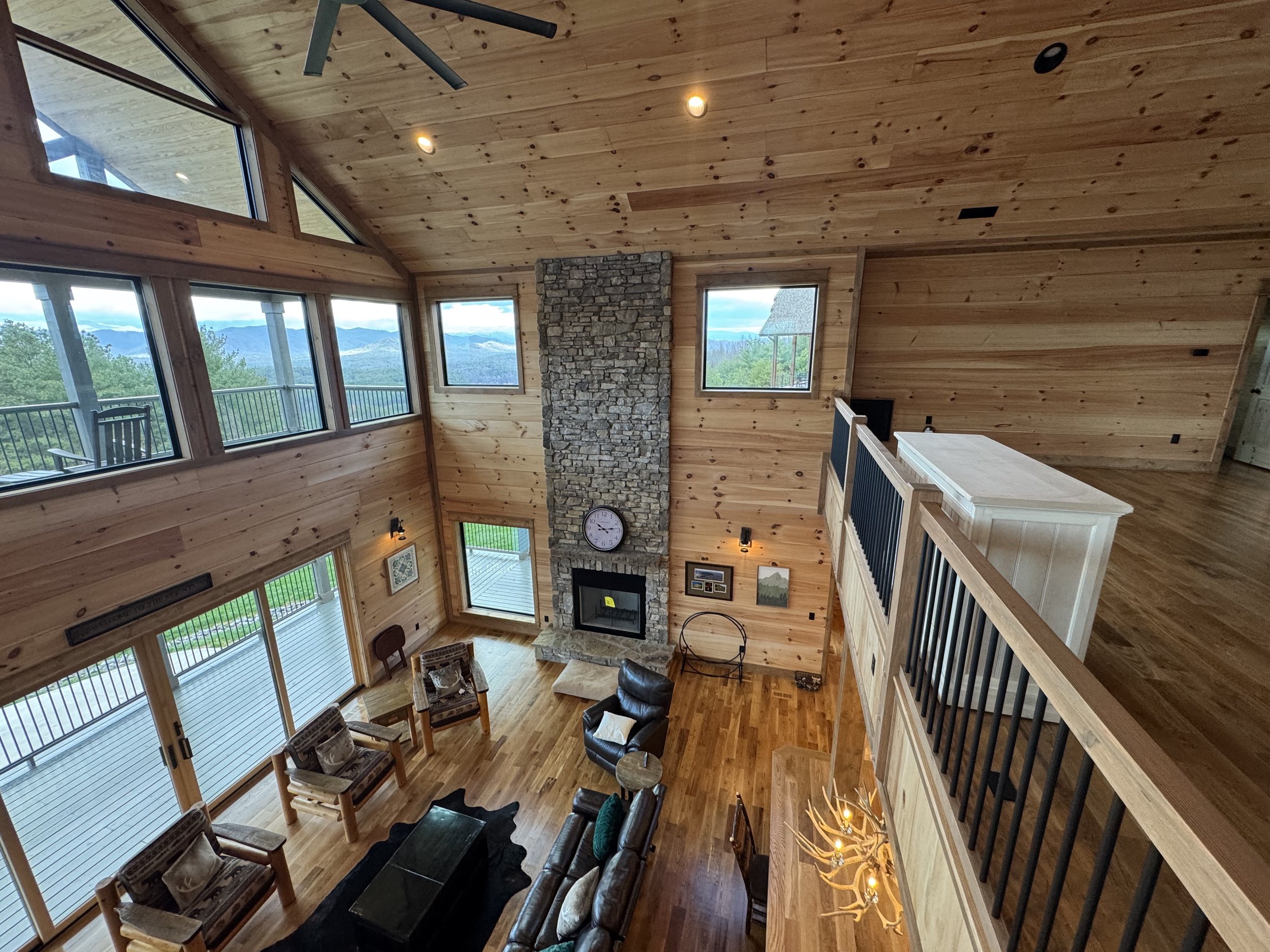 Interior of a spacious loft-style living room with large windows, wooden walls and ceiling, a stone fireplace, and mountain views outside.