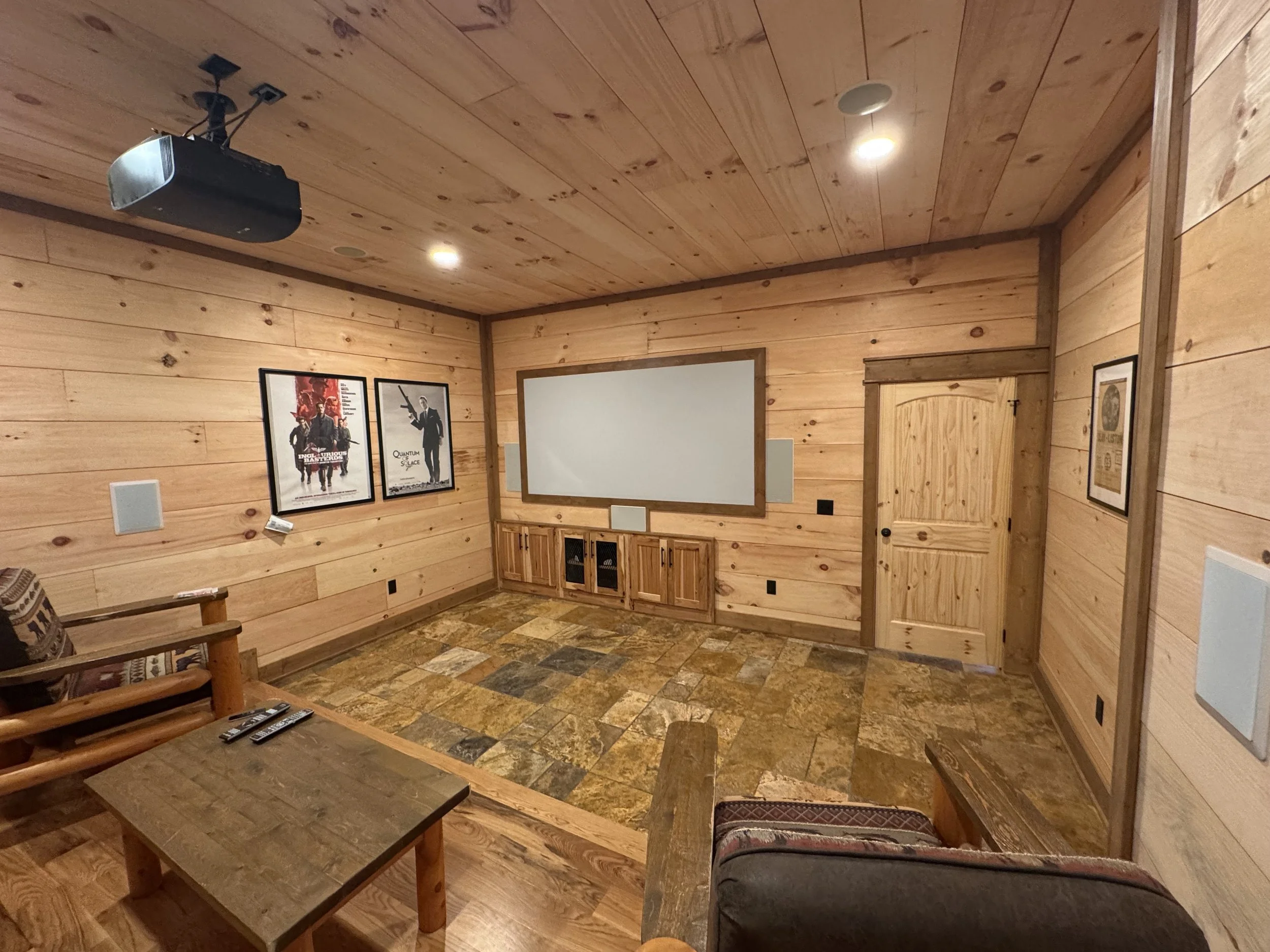 Cozy home theater room with wood-paneled walls and ceiling, movie posters on the wall, a projector mounted on the ceiling, a whiteboard on the wall, and a seating area with a couch and armchair.