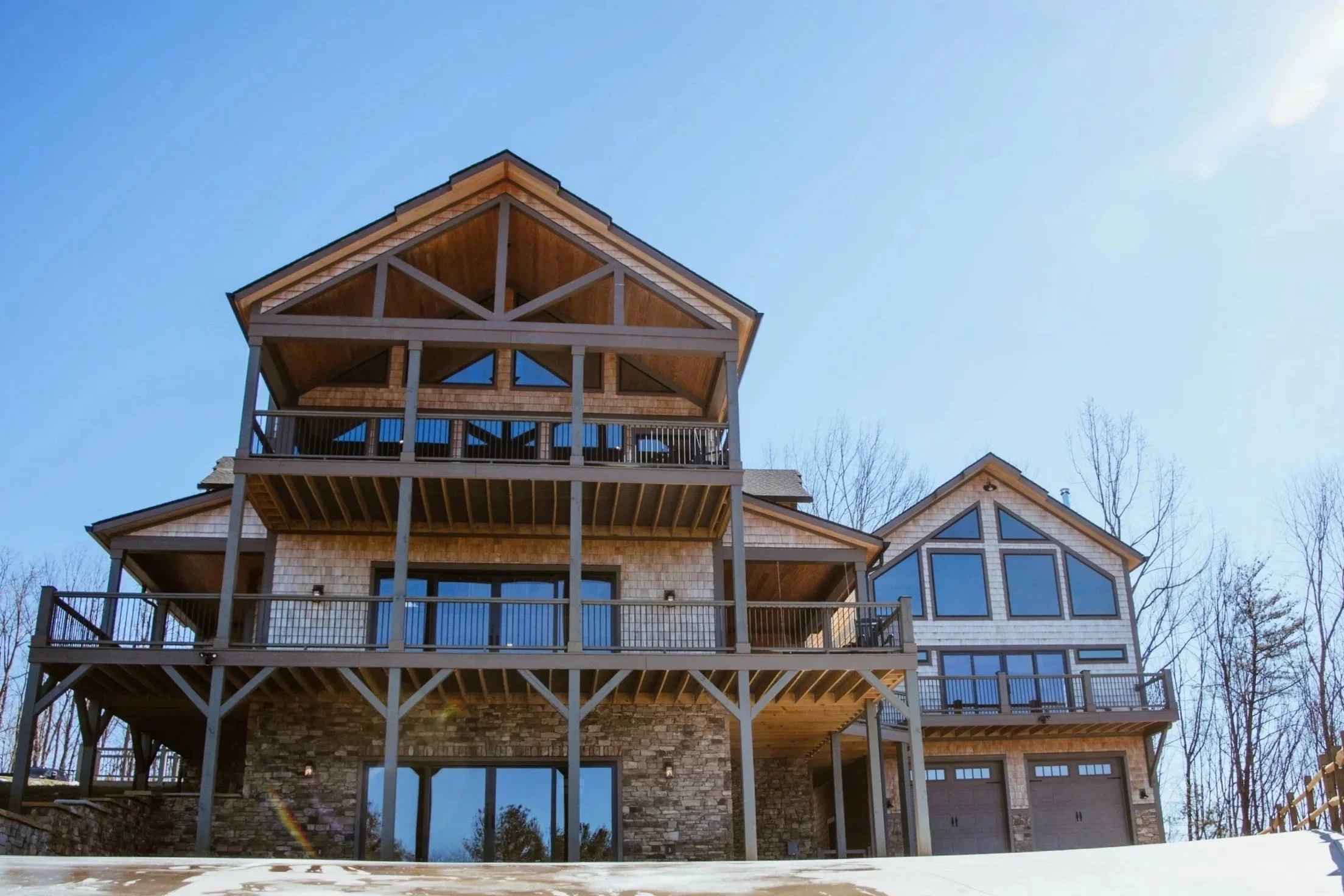 A large modern house with multiple decks, stone and wood exterior, surrounded by leafless trees under a clear blue sky.