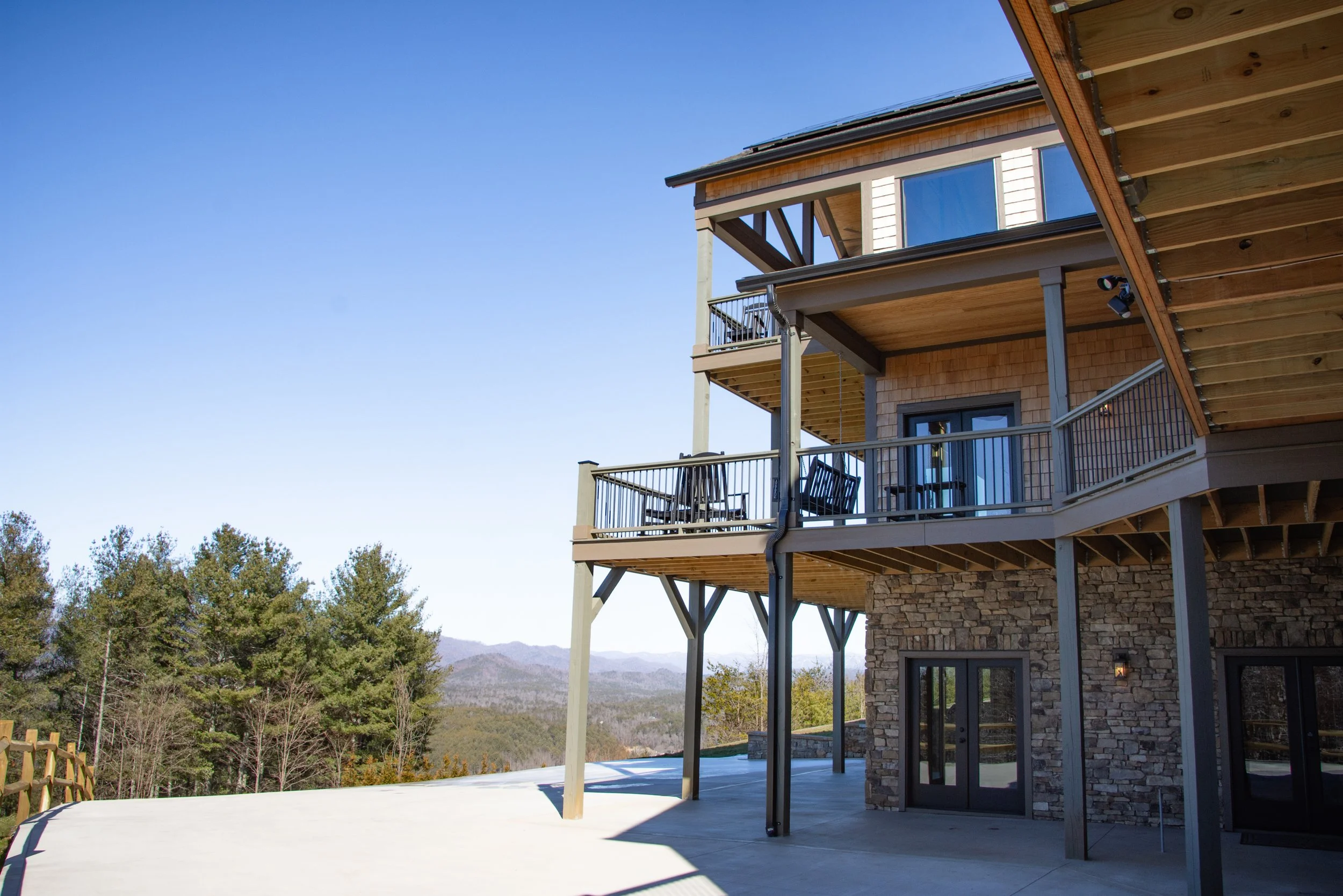 Multi-story house with wooden and stone exterior, multiple balconies with black railings, glass doors, overlooking a scenic mountain landscape, under a clear blue sky.