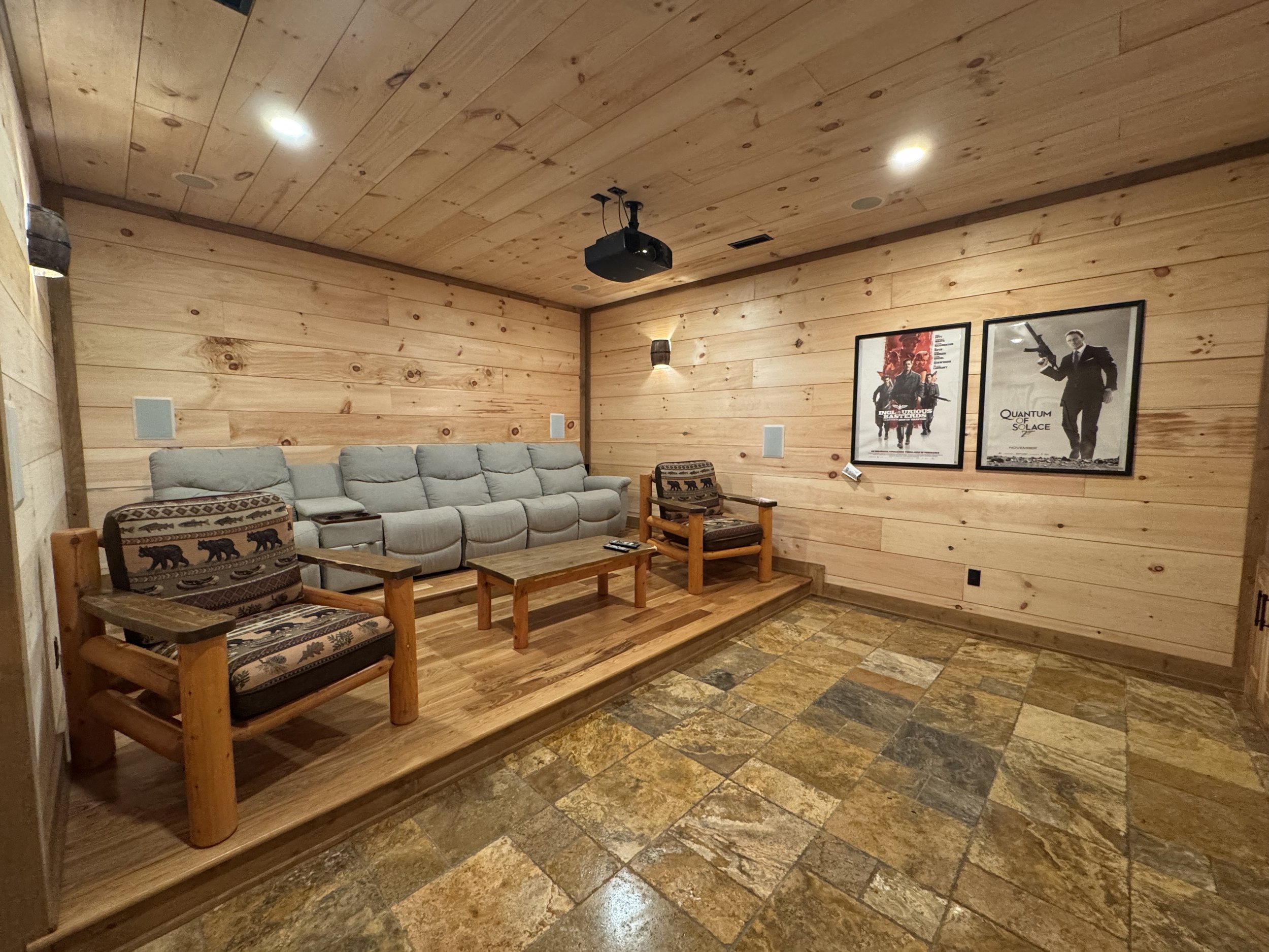 A cozy home theater room with wooden walls and ceiling, featuring a beige couch, two rustic armchairs, a wooden coffee table, framed movie posters, and ambient lighting.
