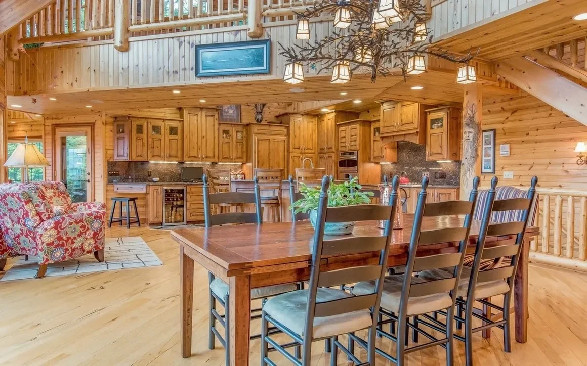 Open-concept kitchen and dining area in a log cabin with wood walls and ceiling, featuring a wooden dining table with eight chairs, a floral-patterned armchair, a small kitchen with wooden cabinets, granite countertops, and a decorative chandelier ha