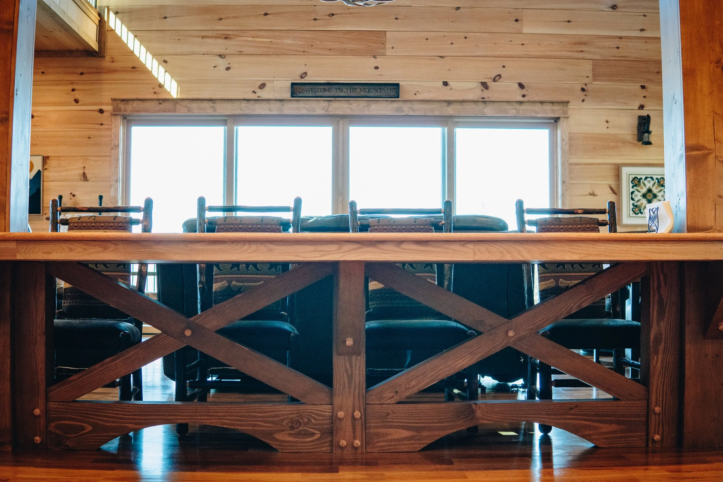 A wooden dining area seen from a lower angle, with a large rectangular table and several chairs. The background features large windows letting in bright light, with wooden walls and a sign that reads 'Welcome to the Mountains'.