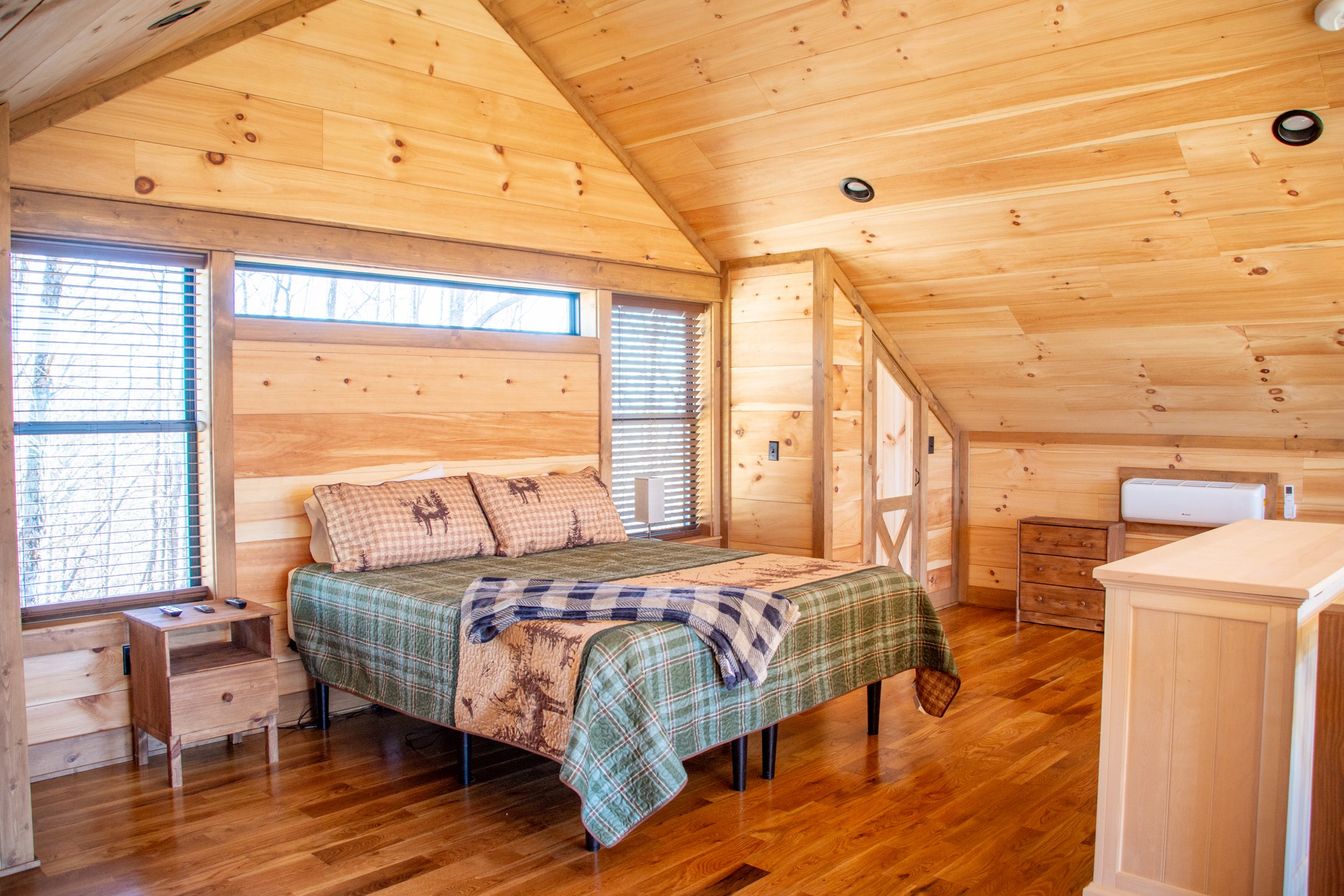 Cozy rustic bedroom with wood-paneled walls and ceiling, a bed with plaid bedding and deer-themed pillows, hardwood floors, a nightstand with remotes, a small dresser, and an air conditioning unit.