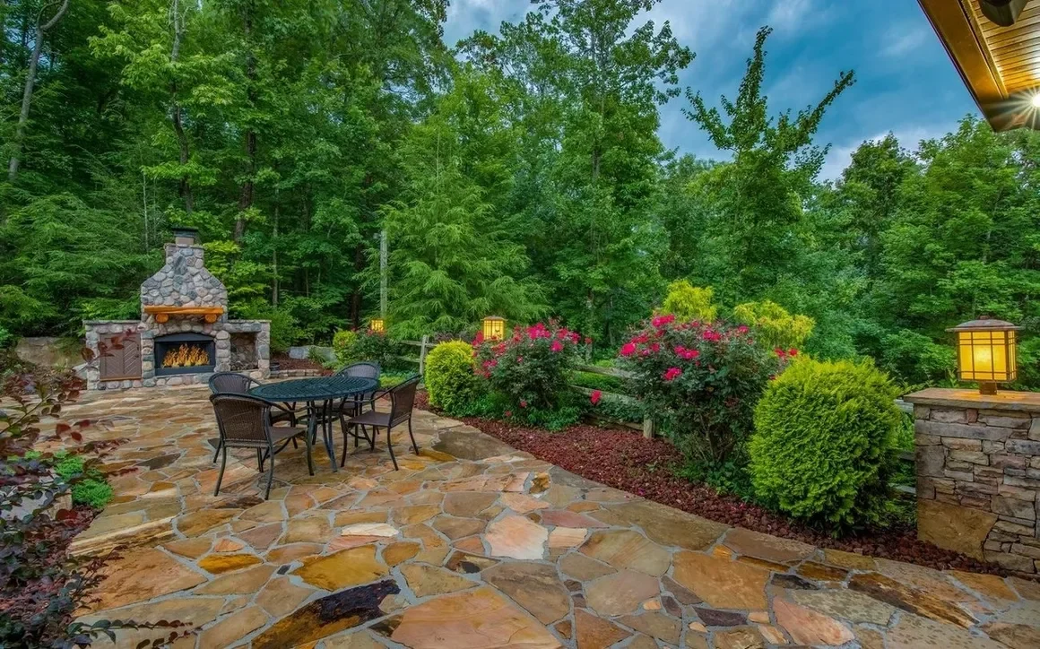 Outdoor patio with a stone floor, surrounded by greenery and colorful bushes, featuring a stone fireplace with a fire burning, a black metal table with four chairs, and lantern-style lights on stone pillars.
