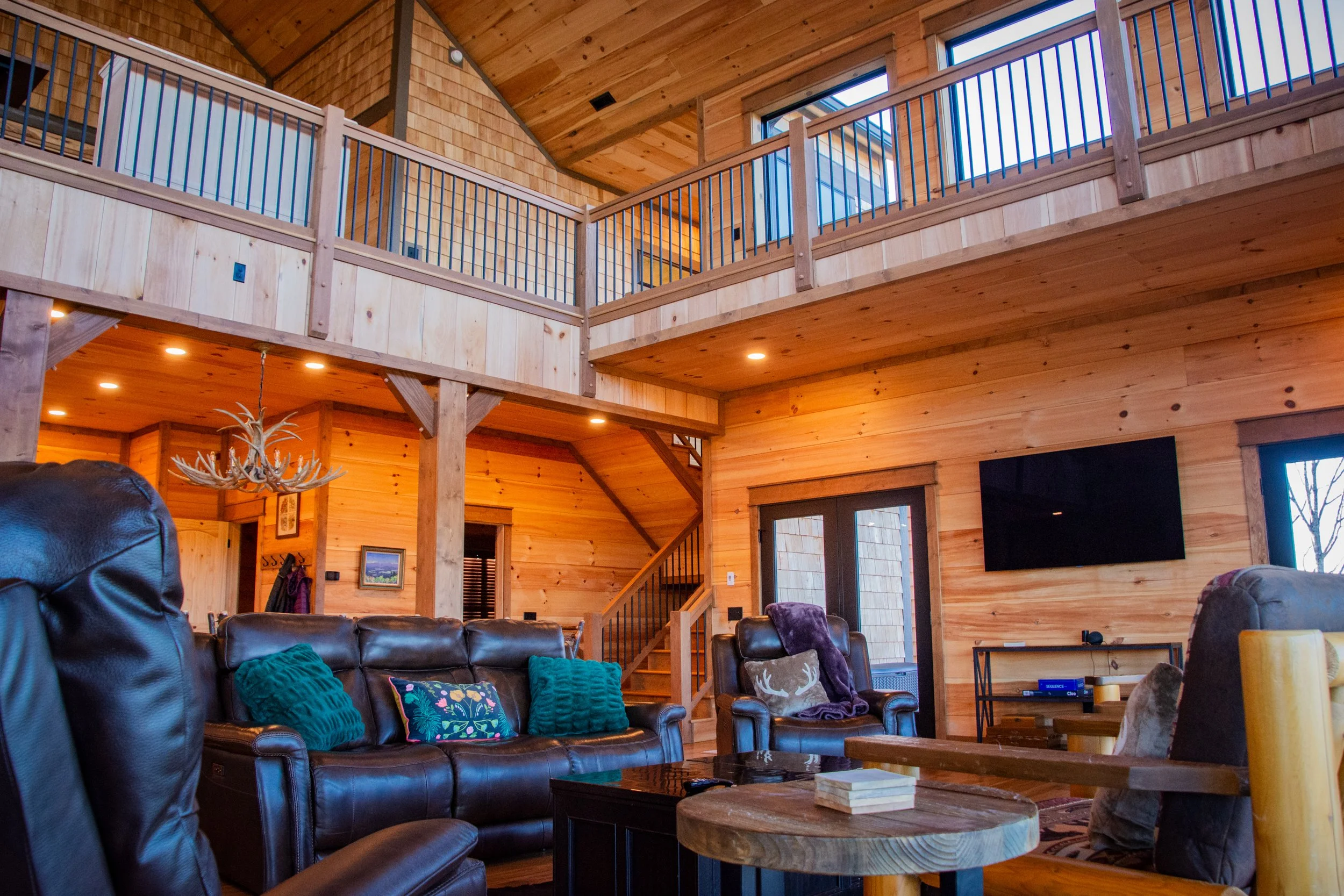 Interior of a wooden cabin living room with leather and fabric armchairs, throw pillows, a wall-mounted TV, staircase, and a second floor balcony with wooden railing.
