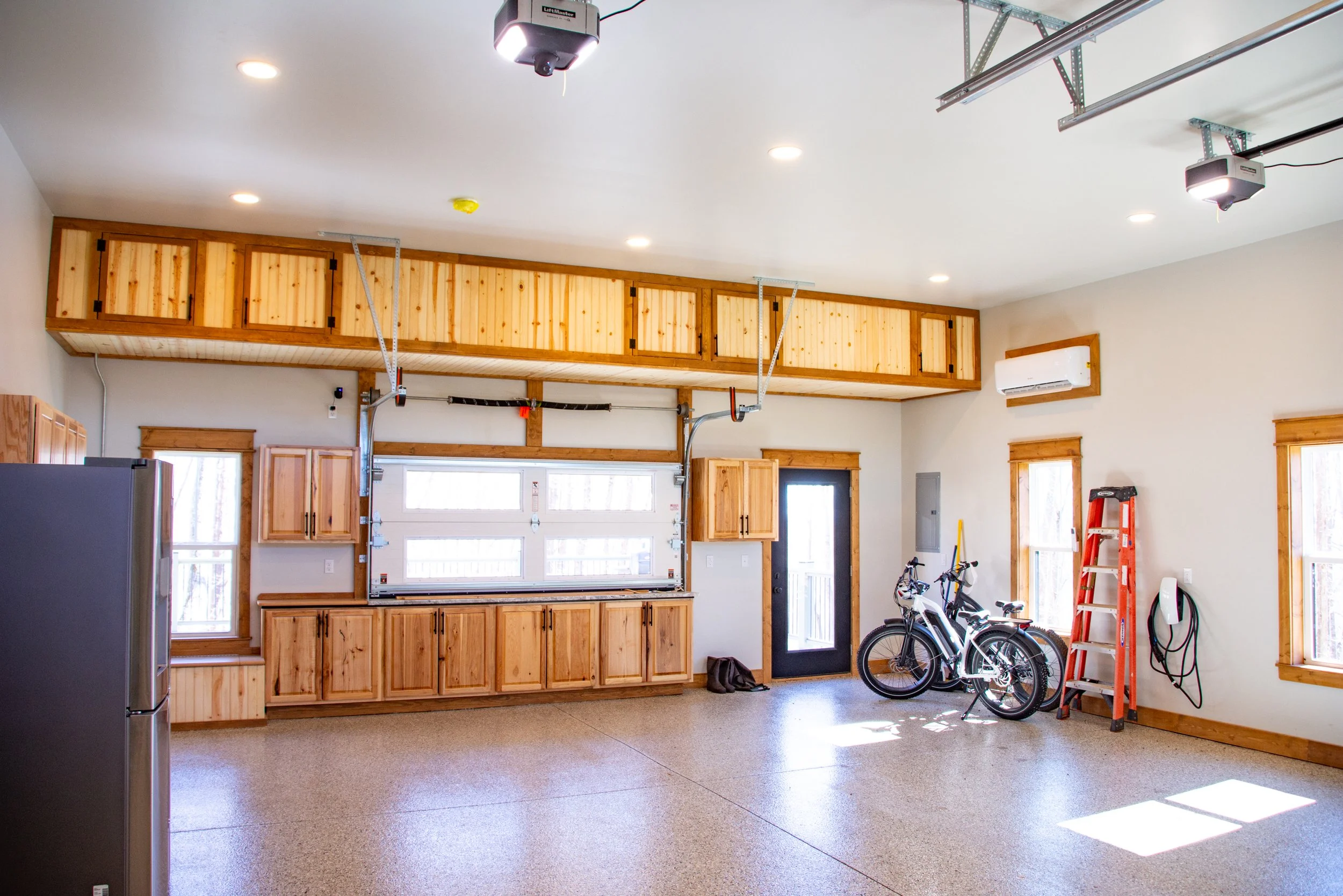 Spacious garage with wooden cabinets, a mountain bike, a red ladder, and a black refrigerator, illuminated by ceiling lights and sunlight through windows.