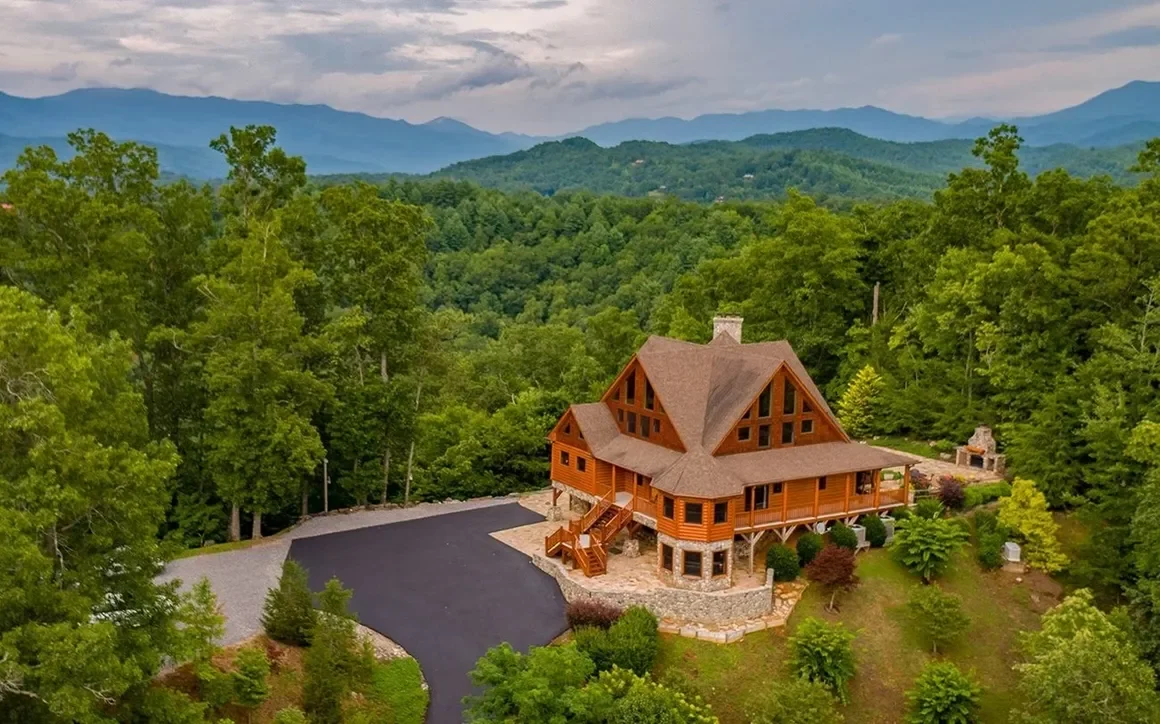 A large wooden house with a steep roof surrounded by green trees in a mountainous area.