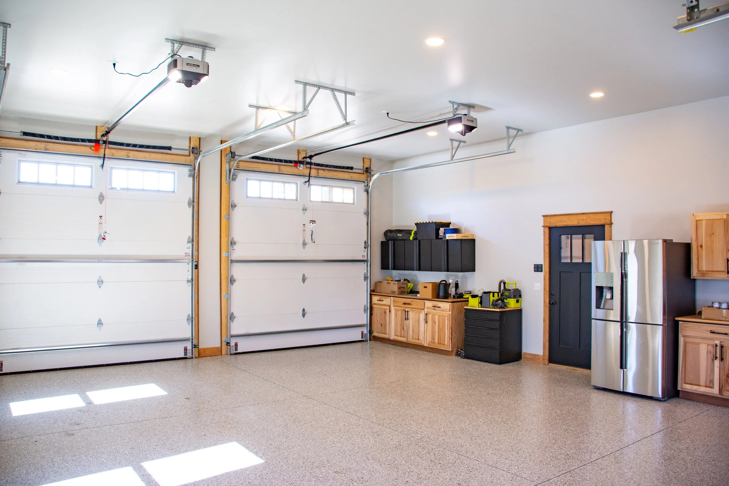 A spacious garage interior with two white sectional garage doors, overhead garage door openers, and windows at the top. Floor is finished with speckled epoxy coating. On the right side, there are black and natural wood storage cabinets, a stainless s
