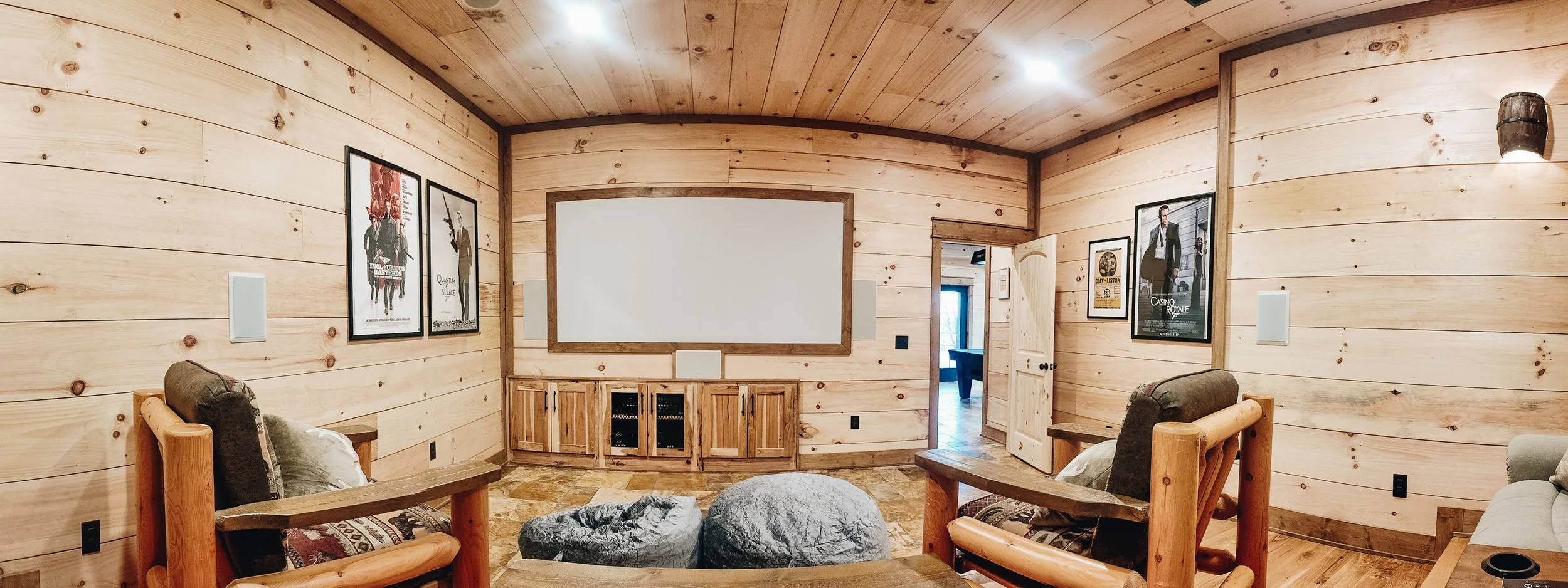 A cozy living room with wooden walls, ceiling, and furniture including chairs and a cabinet. There are framed posters on the walls and a large white screen or panel on the wall. The room appears to be part of a cabin or rustic home.