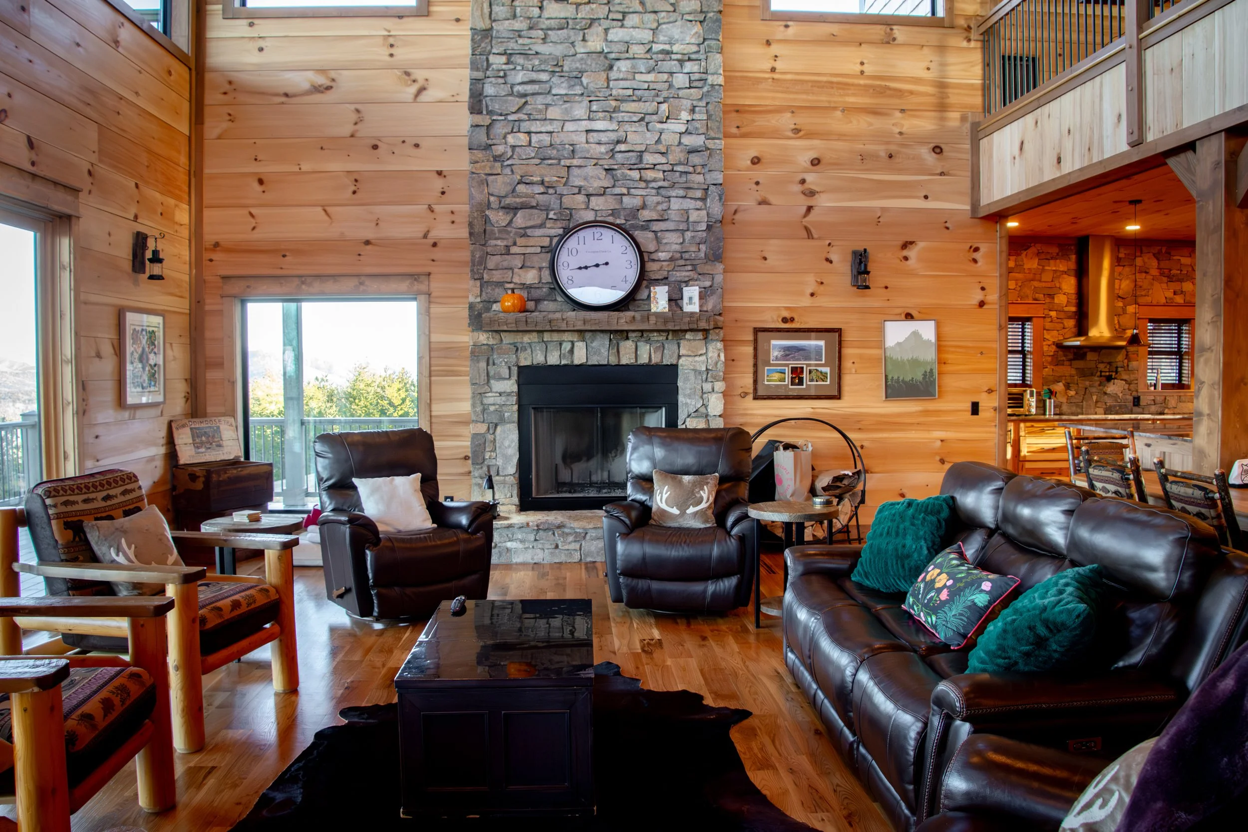 Cozy living room with wooden walls, stone fireplace, leather sofa and chairs, colorful pillows, and framed pictures. Large windows show outdoor trees, a clock hangs above the fireplace, and a black rug lies on the wooden floor.