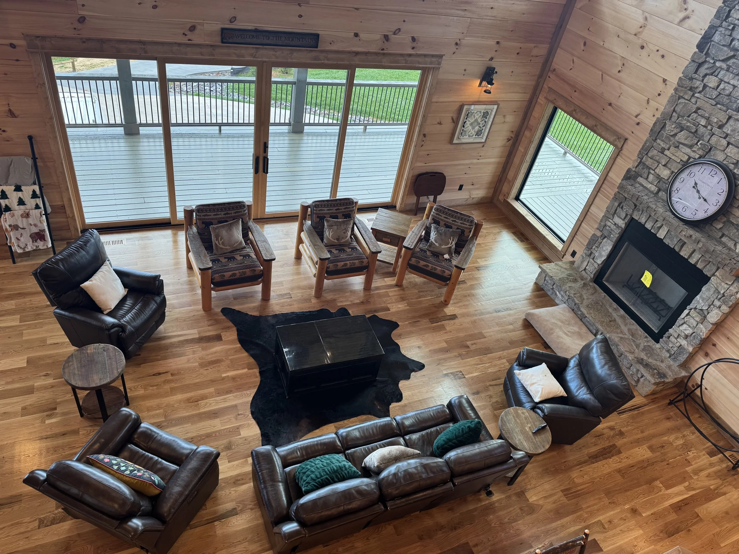 Top-down view of a cozy living room with wooden walls and floor, featuring leather sofas, armchairs, a black coffee table on a black cowhide rug, a stone fireplace with a large round clock above it, sliding glass doors leading to an outdoor deck, and