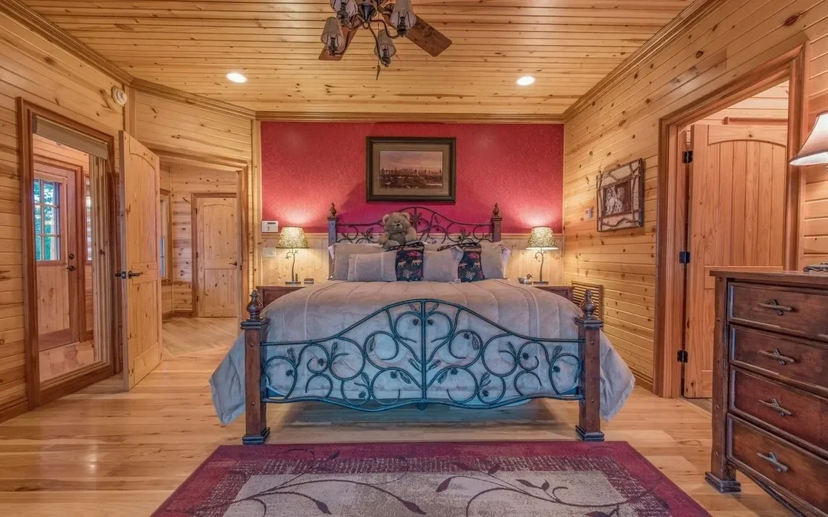 Wood-paneled bedroom with a wrought iron bed, teddy bear, lamps, dresser, mirror, and framed artwork, in a cozy rustic style.