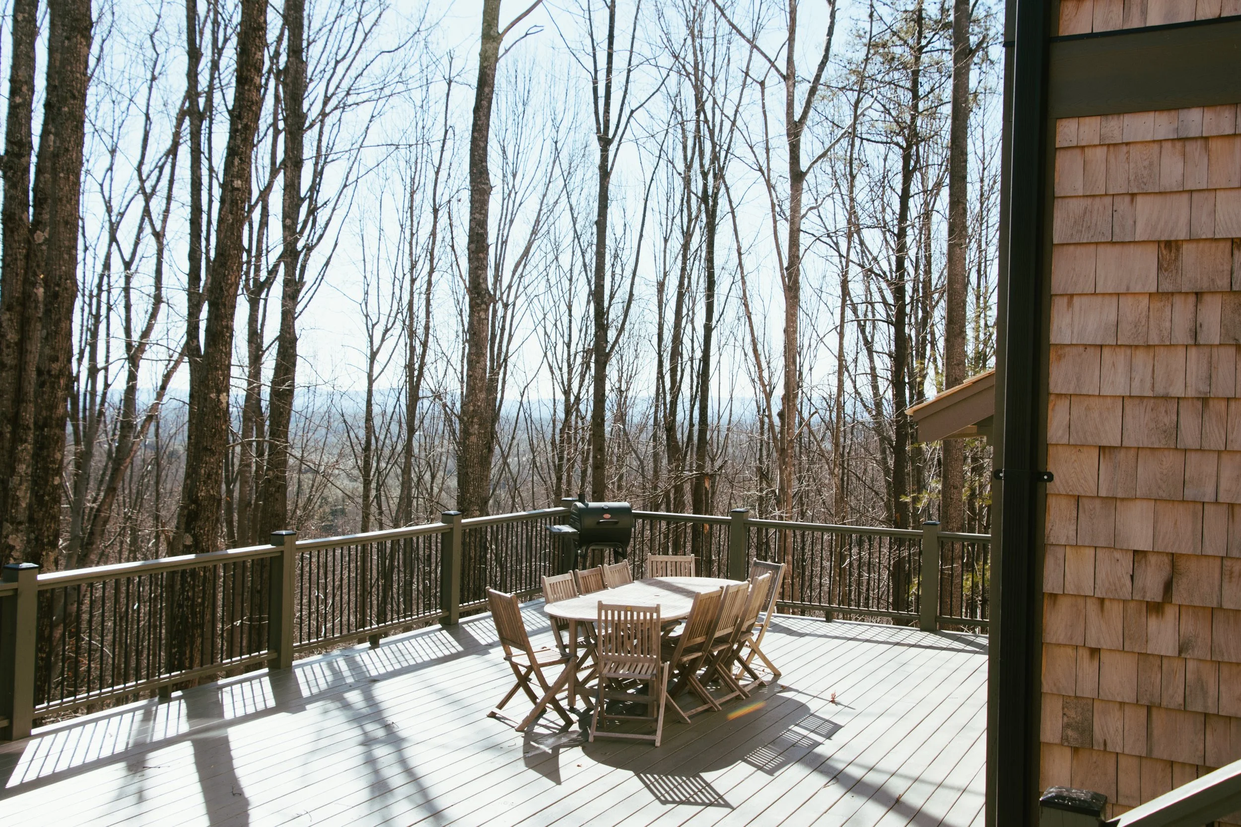 Wooden deck with table and chairs, fenced, overlooking leafless trees in a wooded area, on a bright, clear day.