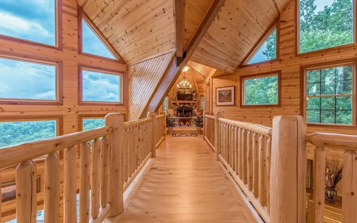 Wood-paneled hallway with a wooden railing, triangular windows, and a view of a house's interior with furniture and a fireplace in the distance.