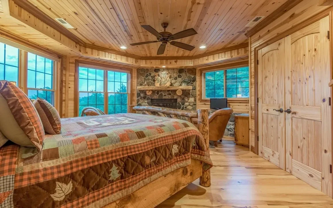 Bedroom with log cabin style wood paneling, large windows, stone fireplace, ceiling fan, and wooden furniture.