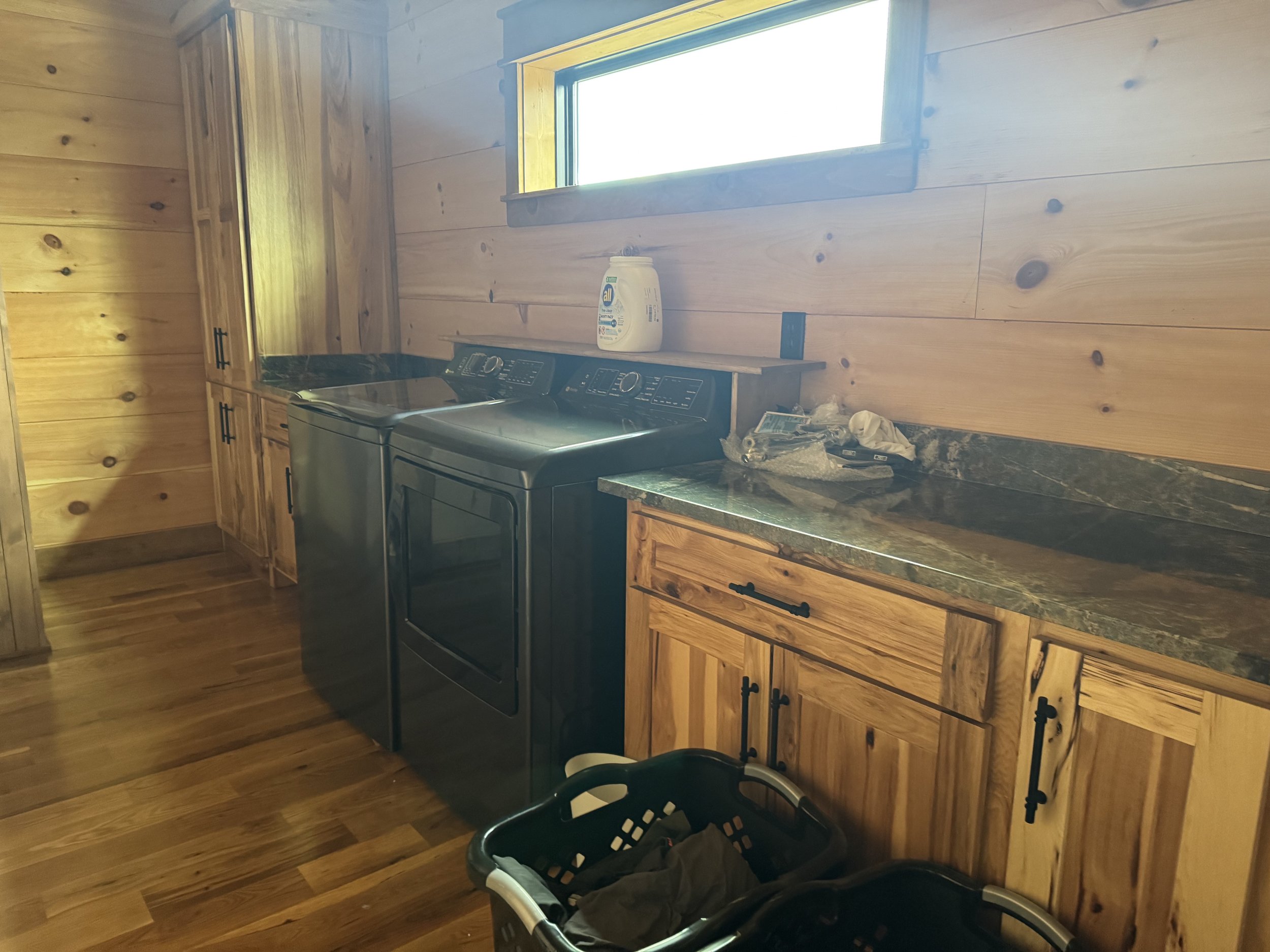 Laundry room with wooden walls, a window, a dark gray washing machine and dryer, and wooden cabinets with black handles. There is a laundry basket with clothes in the foreground.