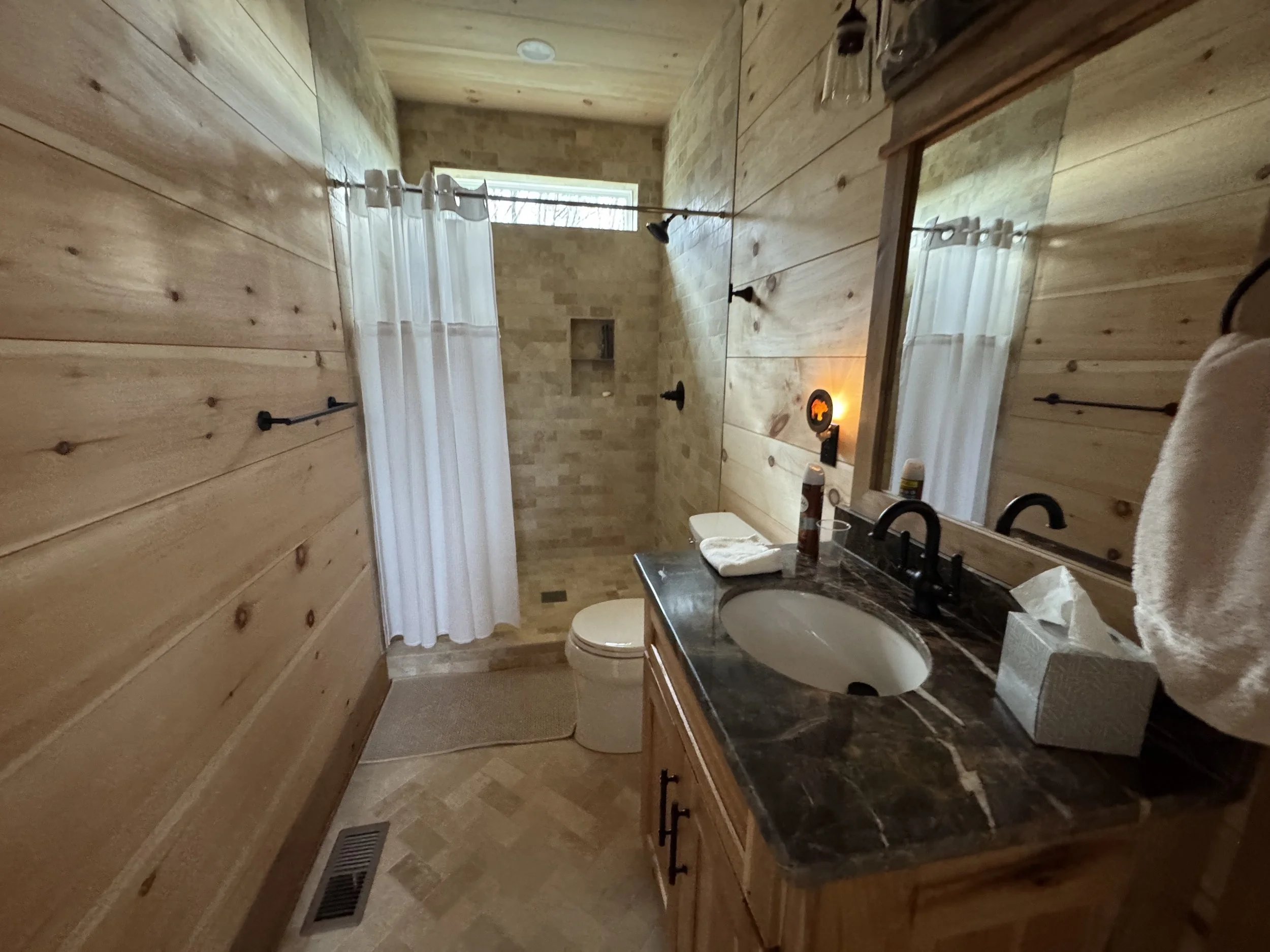 A rustic bathroom with wood-paneled walls, a dark marble countertop with an oval sink, a large mirror, a towel hanging on the right, toiletries on the counter, and a walk-in shower with a curtain and a window above it.