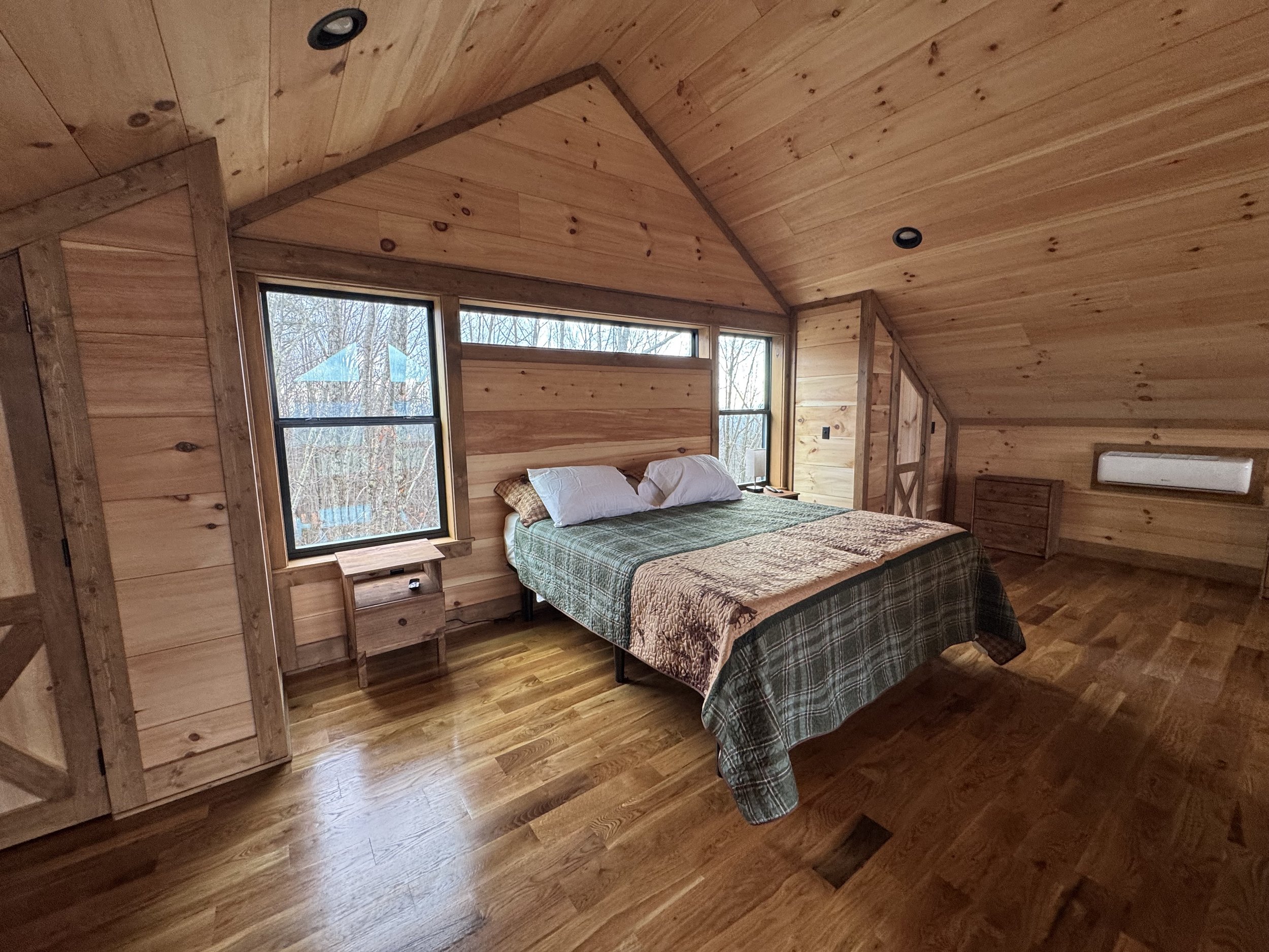 A cozy bedroom with wooden walls, ceiling, and floor, featuring a double bed with pillows and a quilt, located by large windows that reveal a view of trees outside.