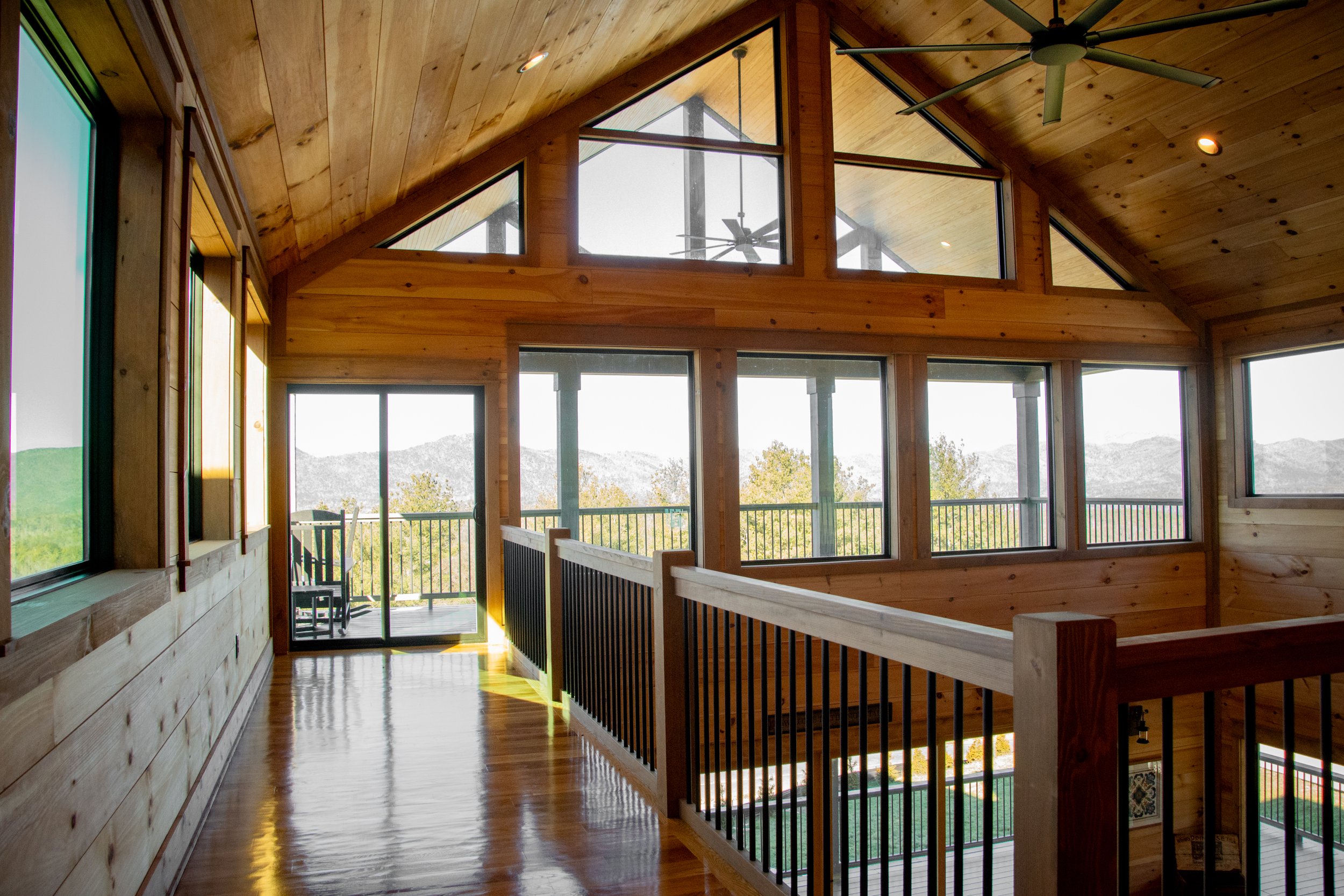 A bright, wooden interior with large windows, a ceiling fan, and a view of mountains and trees outside. There is a wooden rail along the staircase.
